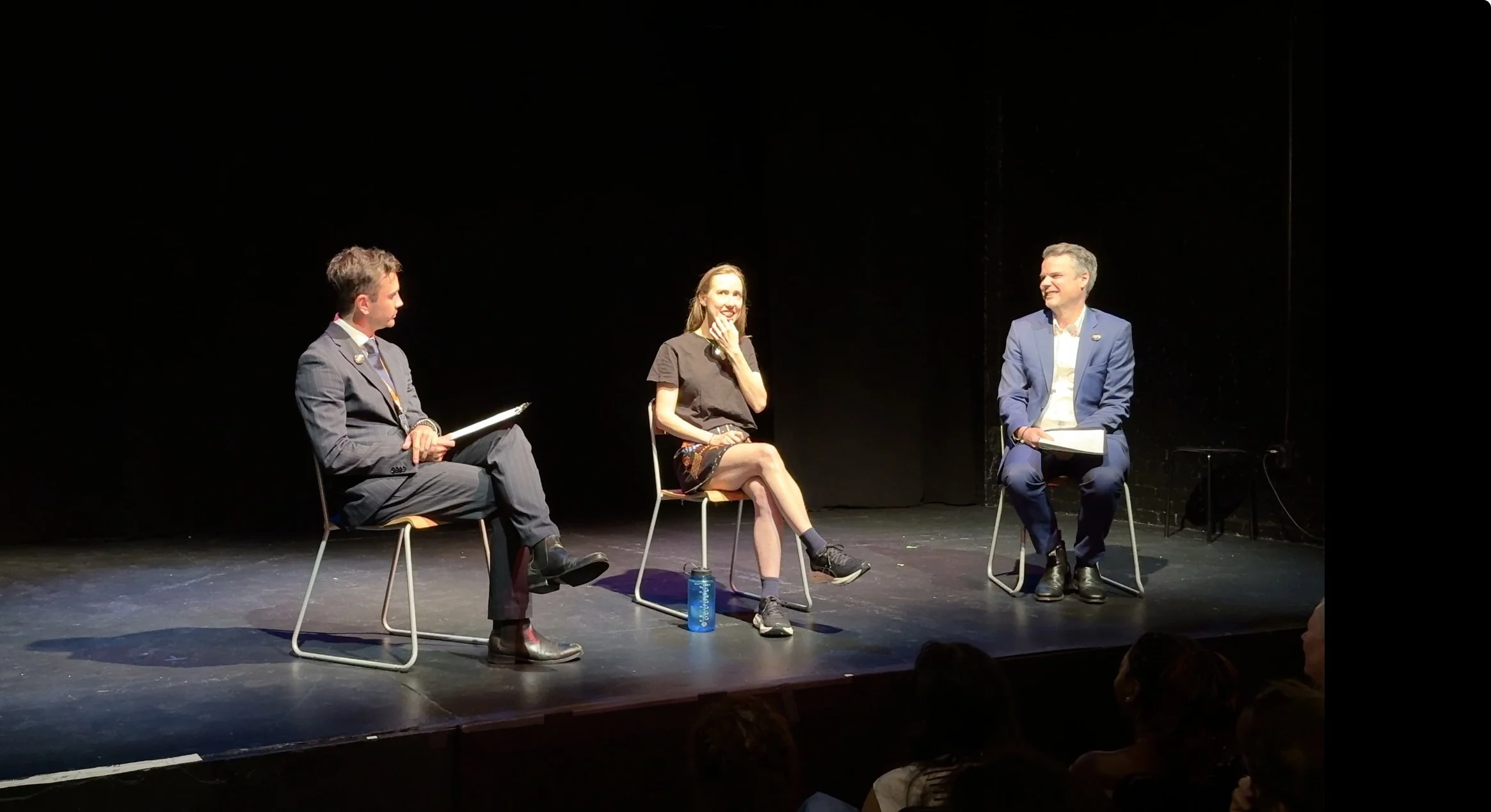 Three people sitting on chairs on a stage in a panel discussion, with a dark background and an audience.