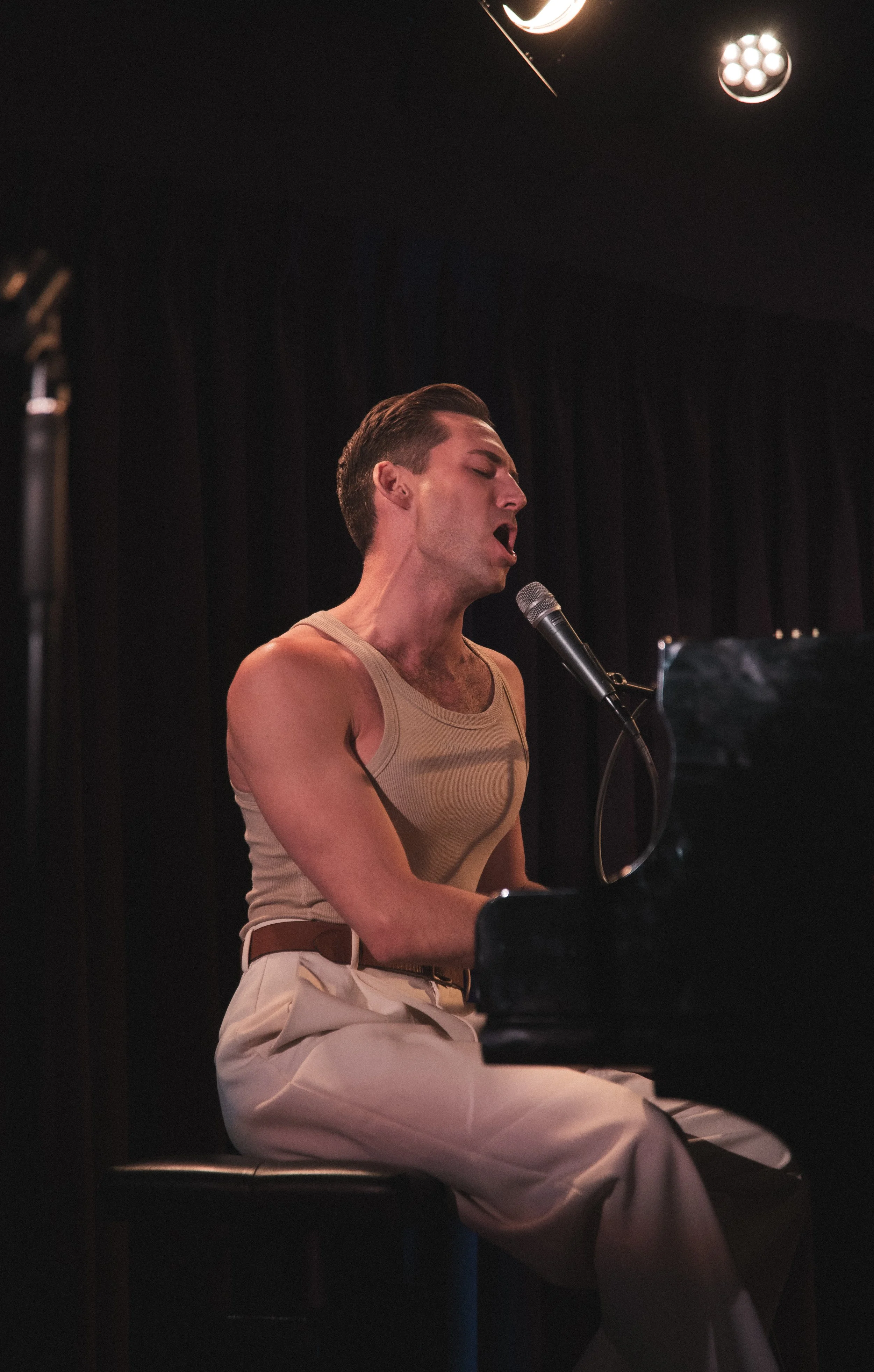 A man passionately singing into a microphone while playing a grand piano in a dimly lit stage setting.
