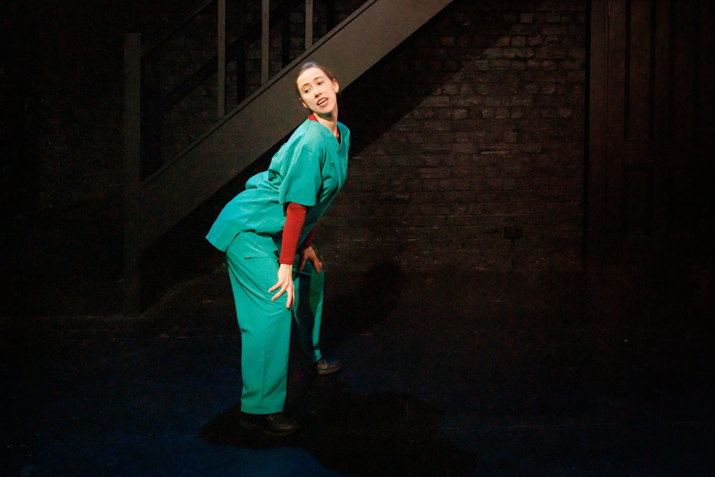 A woman in a teal medical uniform with a red long sleeve shirt underneath stands on a dark stage near a black brick wall and a staircase, performing in a theatrical production.