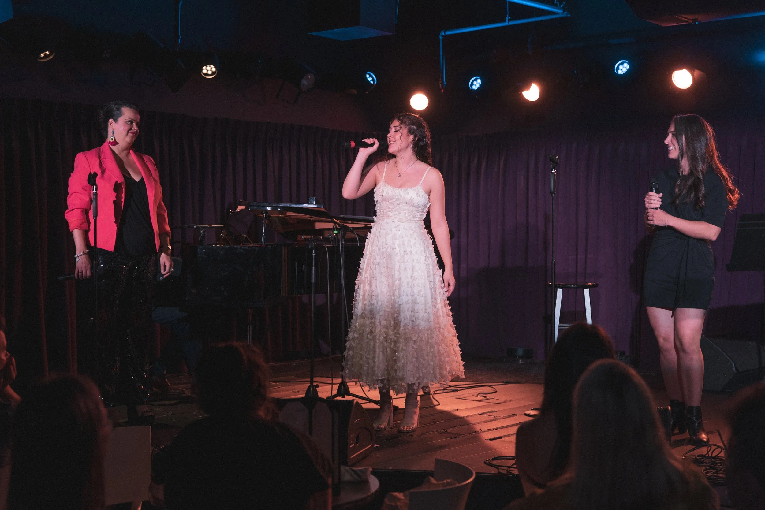 Three women performing on stage in a nightclub, with a grand piano and microphones, under stage lights, and audience in foreground.