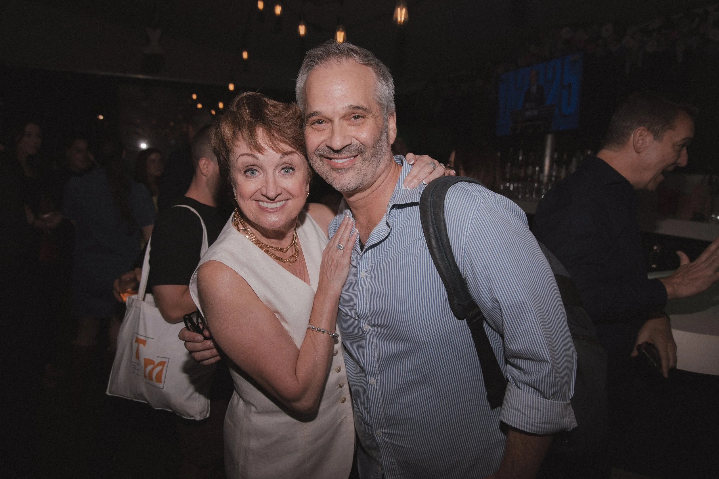 A smiling woman with short brown hair and a pearl necklace, posing with a man with gray hair and a beard, wearing a striped blue shirt, in a dimly lit social event or nightclub.