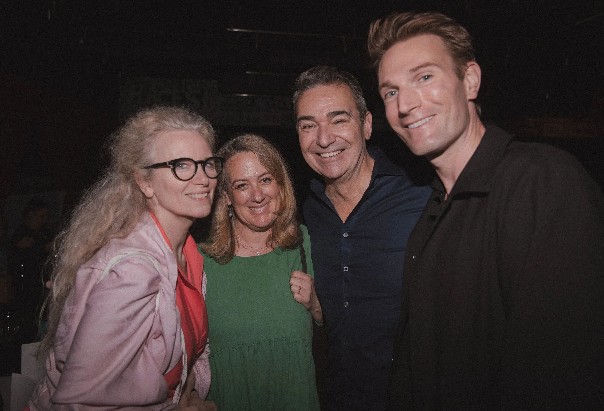 Four smiling people at an indoor event, two women and two men, standing close together.