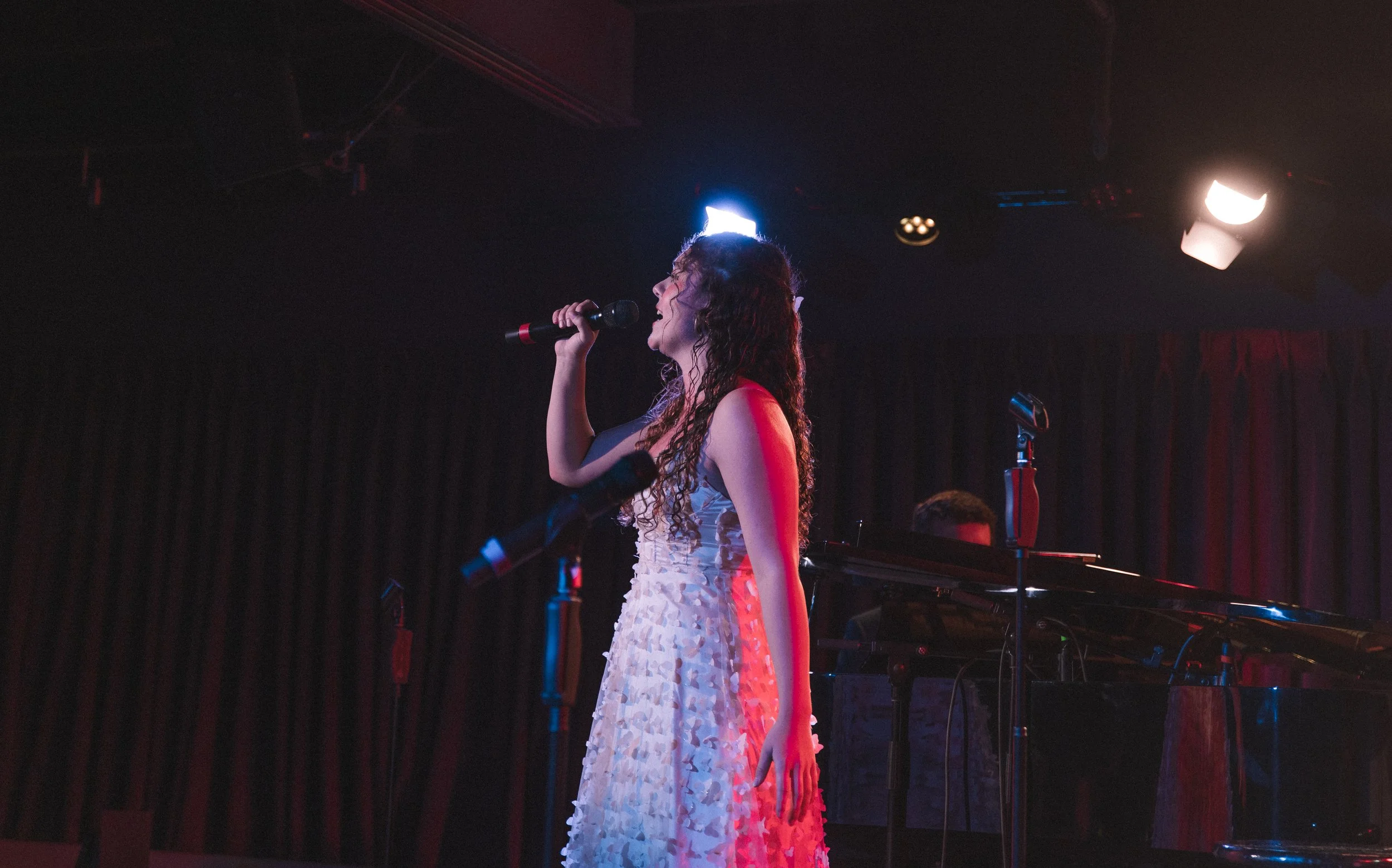 A woman singing into a microphone on stage with a pianist and stage lights in the background.
