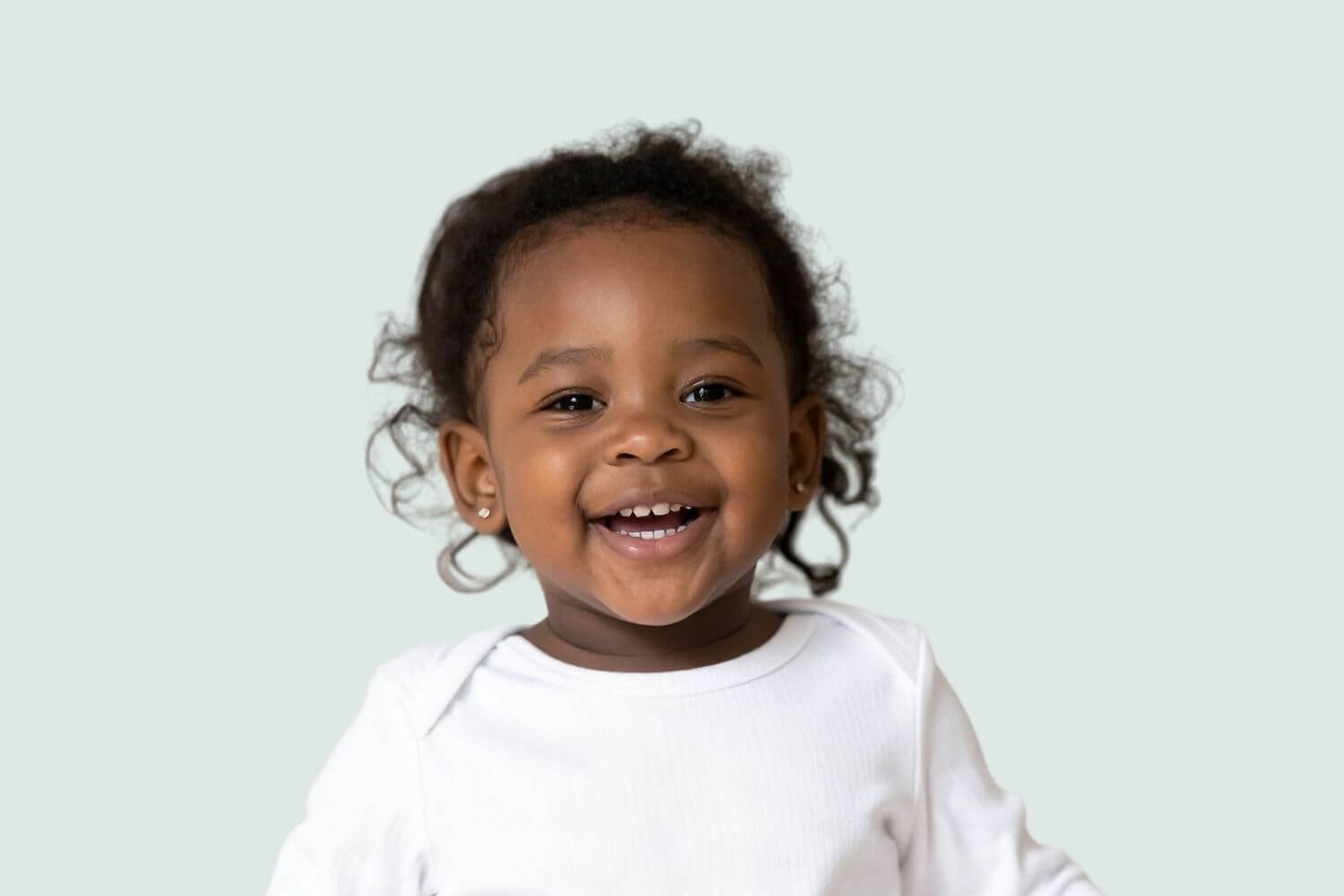 Toddler girl with dark hair smiling wearing a white shirt and earrings.