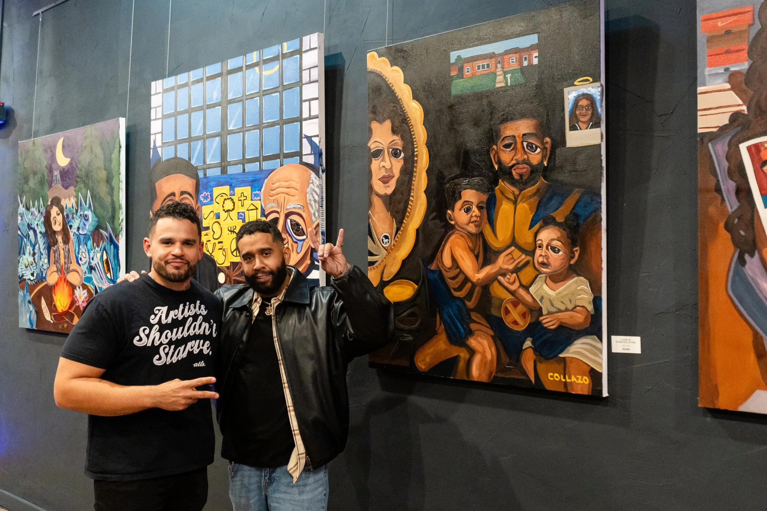 Two men standing in front of colorful paintings in an art gallery, one making a peace sign with his hand.
