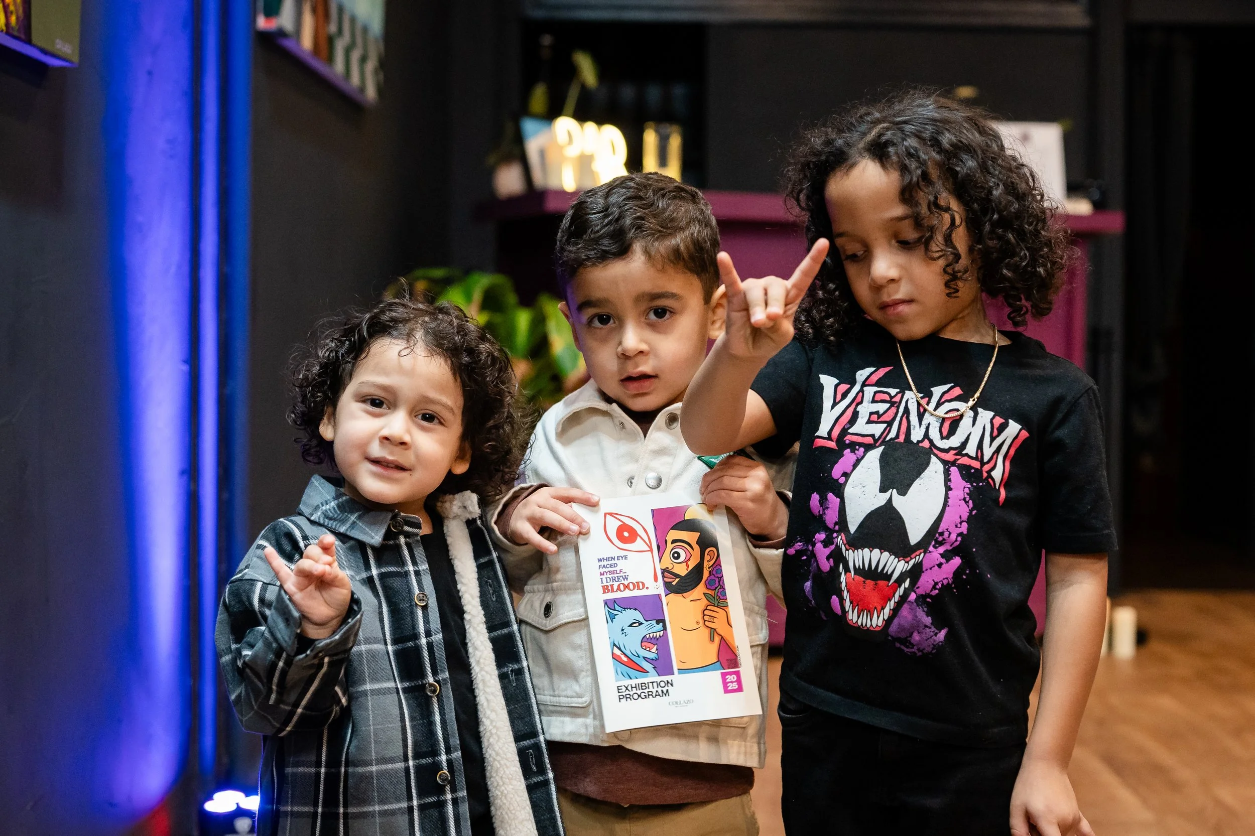 Three children standing indoors, holding a colorful booklet, with one making a rock-and-roll hand gesture.
