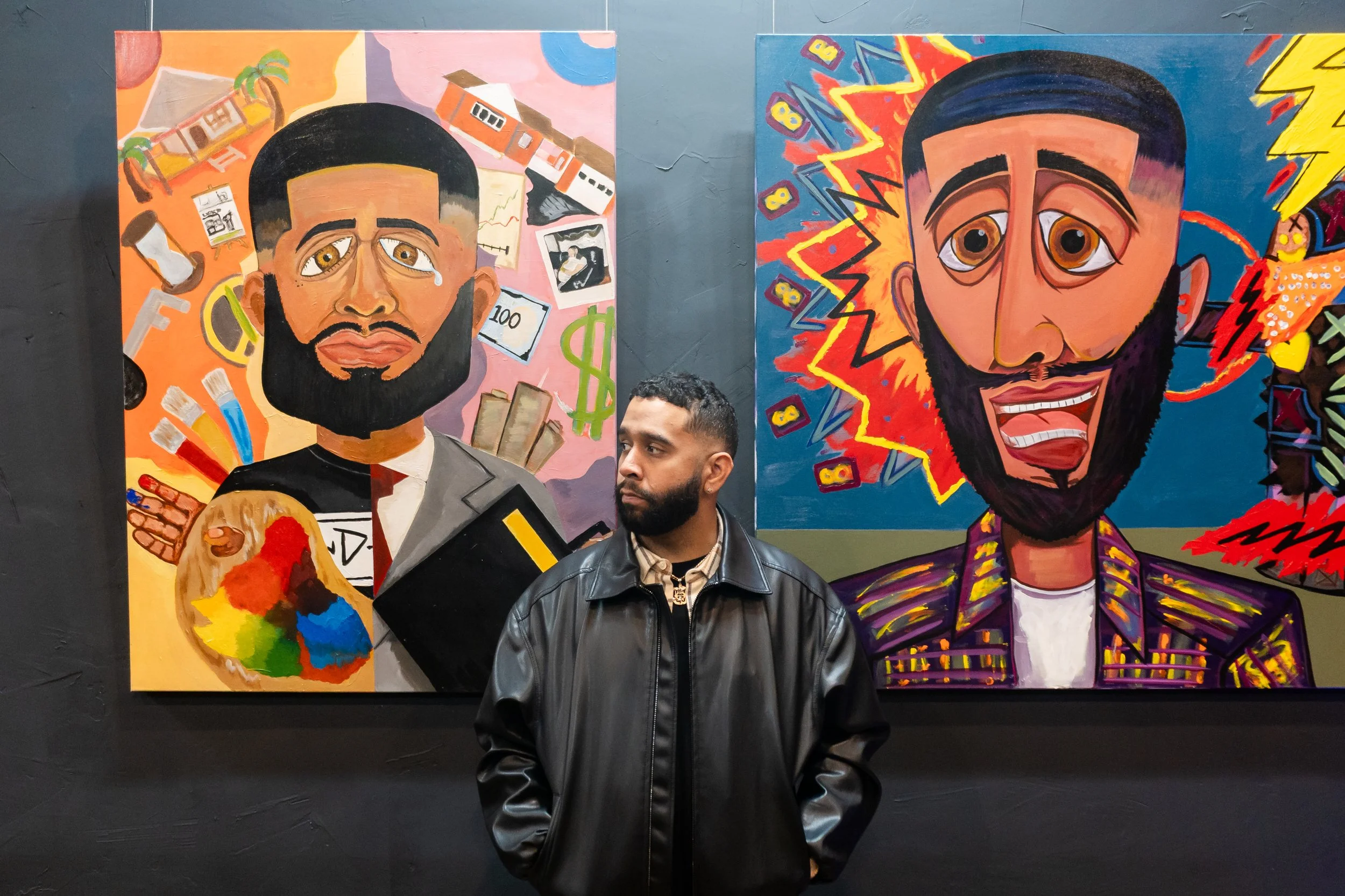 A man with a beard and black leather jacket standing in front of two colorful, abstract portrait paintings of men with beards displayed on a dark wall.