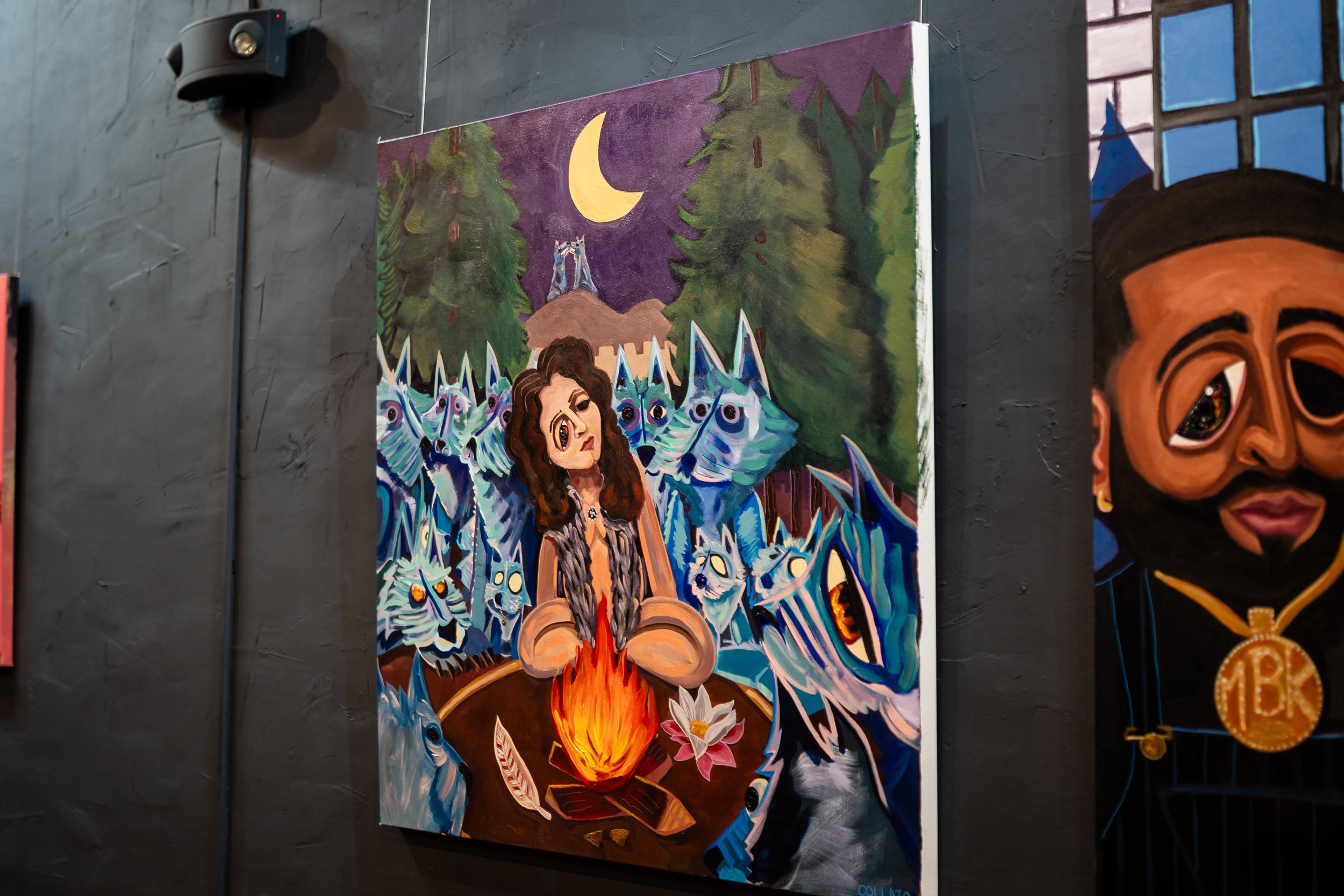 A colorful painting of a woman sitting at a table with a fire in the center, surrounded by blue dragon-like creatures with large eyes, set against a forest background at night with a moon in the sky.