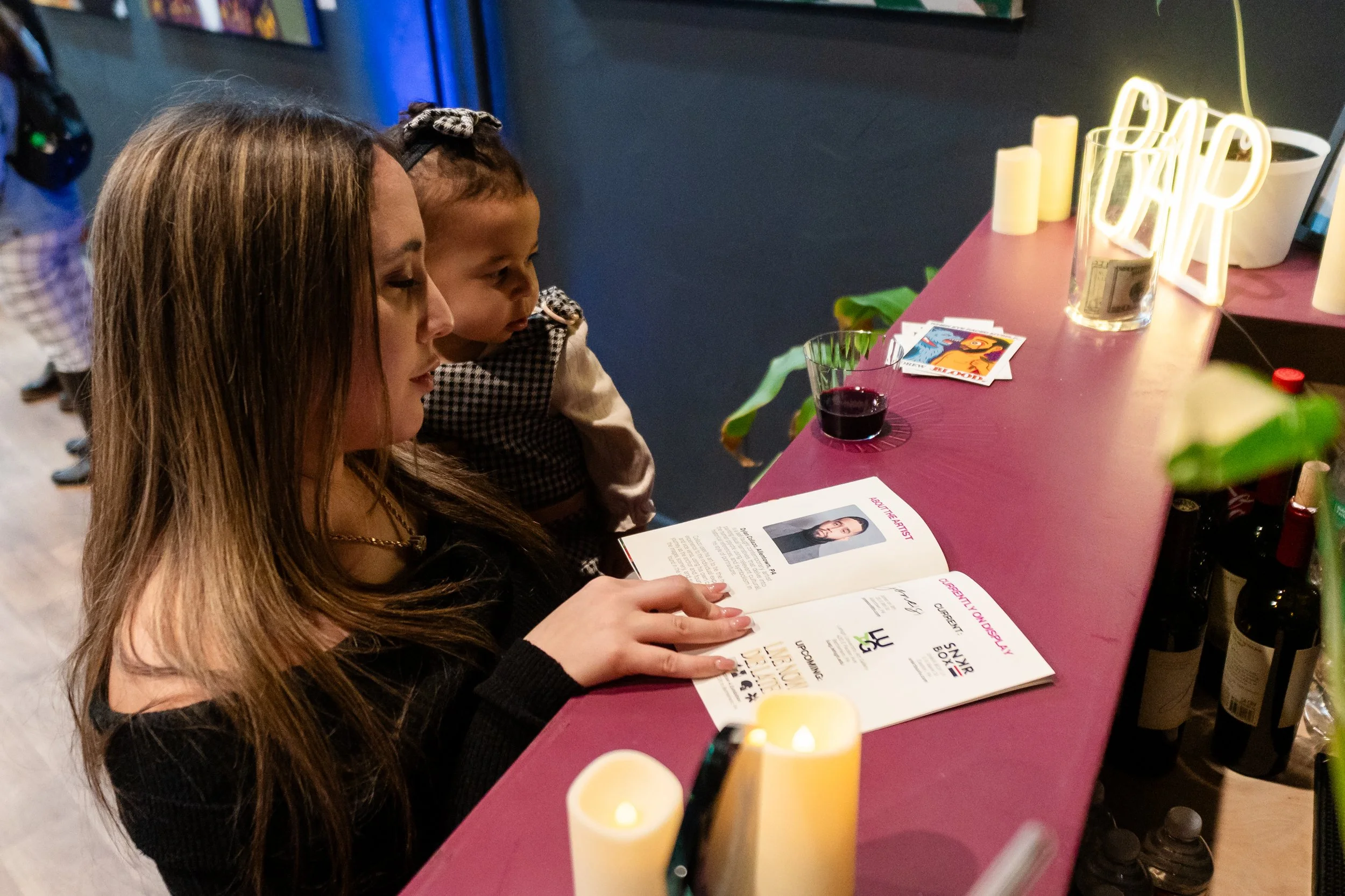 A woman and young girl reading a program or brochure at a bar or cafe counter with candles, a drink, and decorative items.