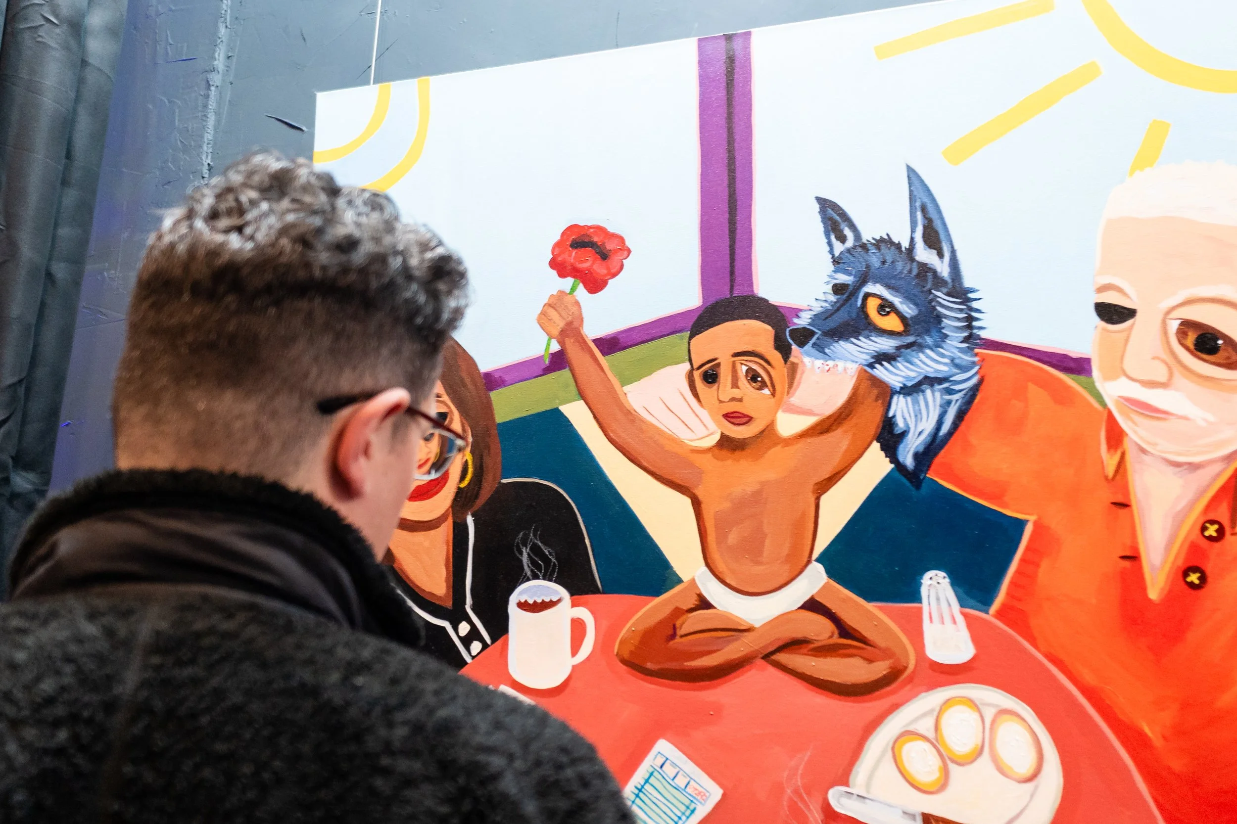 A person with glasses is looking at a colorful painting depicting a child with dark skin, sitting cross-legged at a table, holding a red flower in one hand and a wolf's head in the other. The painting includes other figures and breakfast items on the