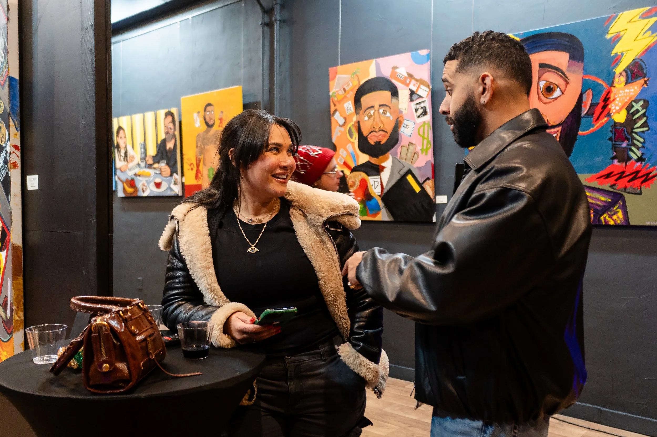 A woman and a man talking at an art gallery, with colorful paintings on the wall behind them.