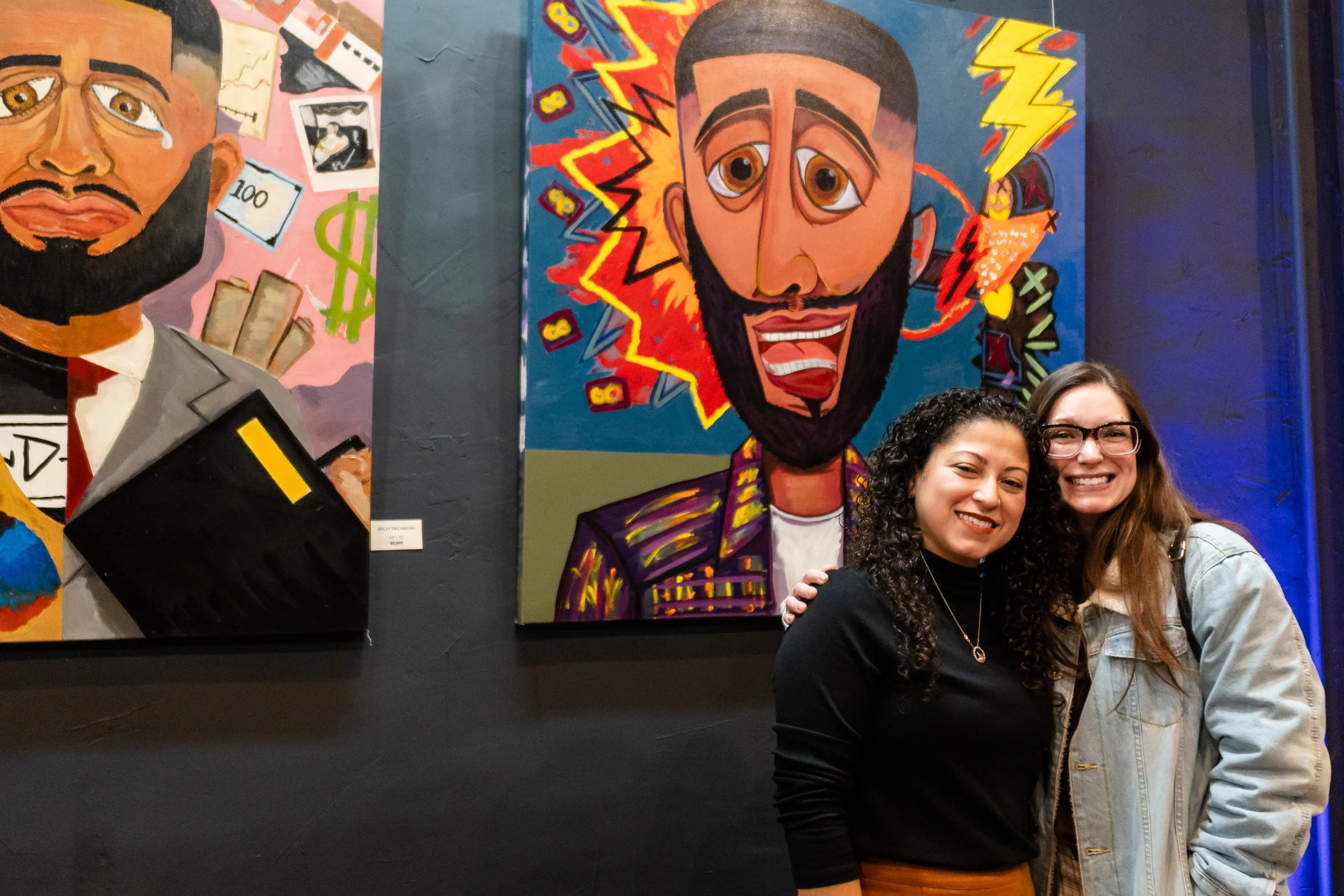 Two women smiling in front of colorful paintings at an art gallery.