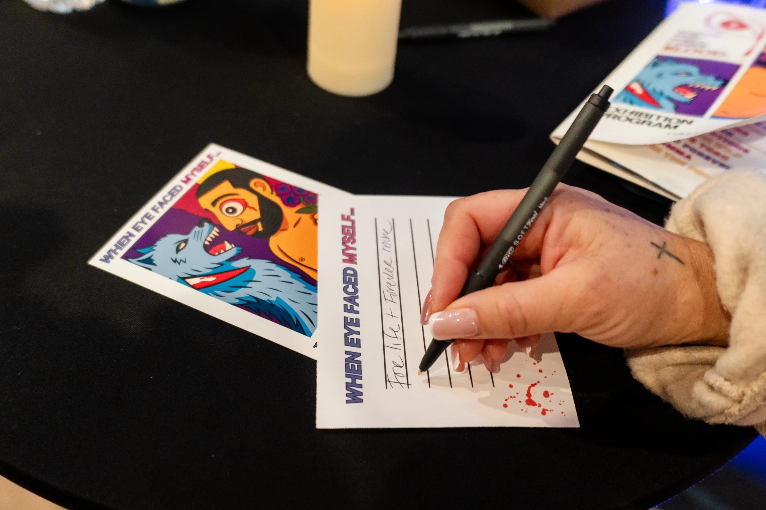 A person writes on a card with a black pen, the card reads 'The AC + FRONTLINE MOVE' and features a colorful illustration of a man with a beard and a wolf. There are other similar cards on a black table.