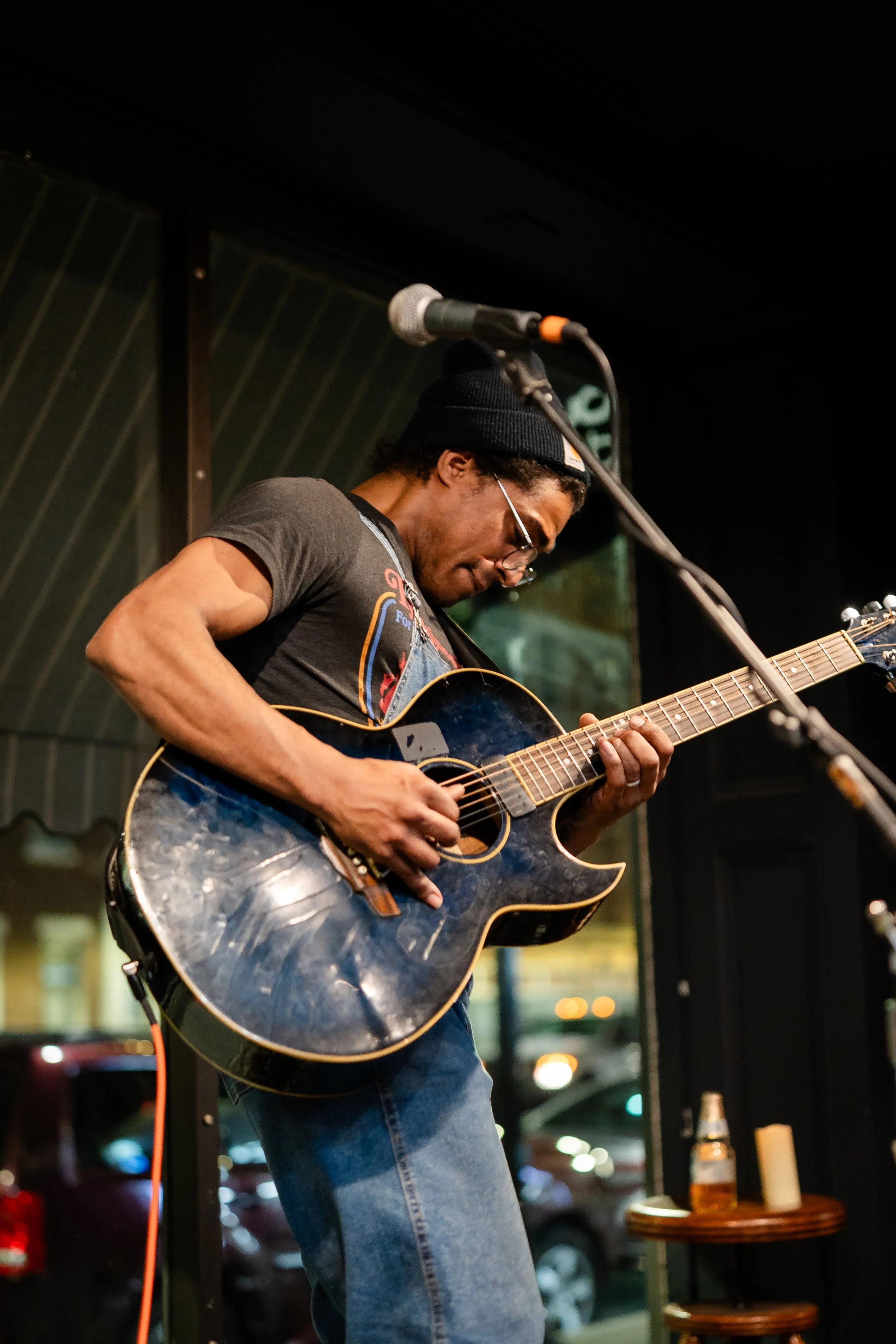 A musician wearing glasses, a beanie, and a black t-shirt is playing an acoustic guitar on stage.