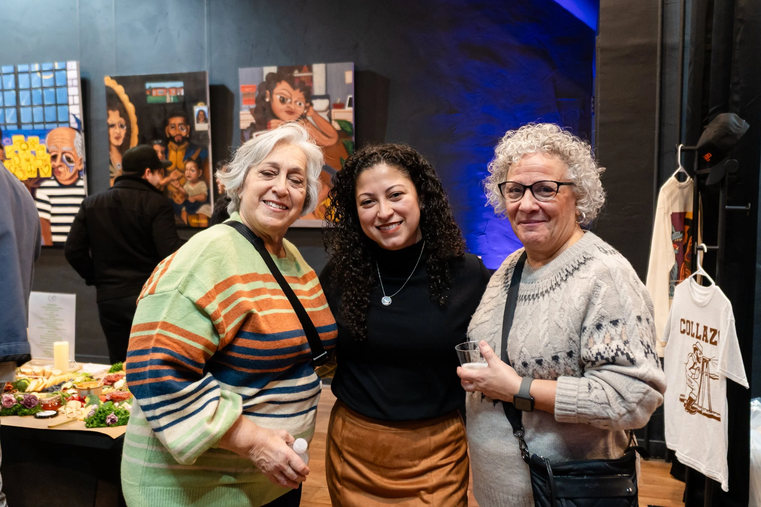 Three women smiling and posing for a photo at an indoor art event, with colorful paintings in the background and a table of food nearby.