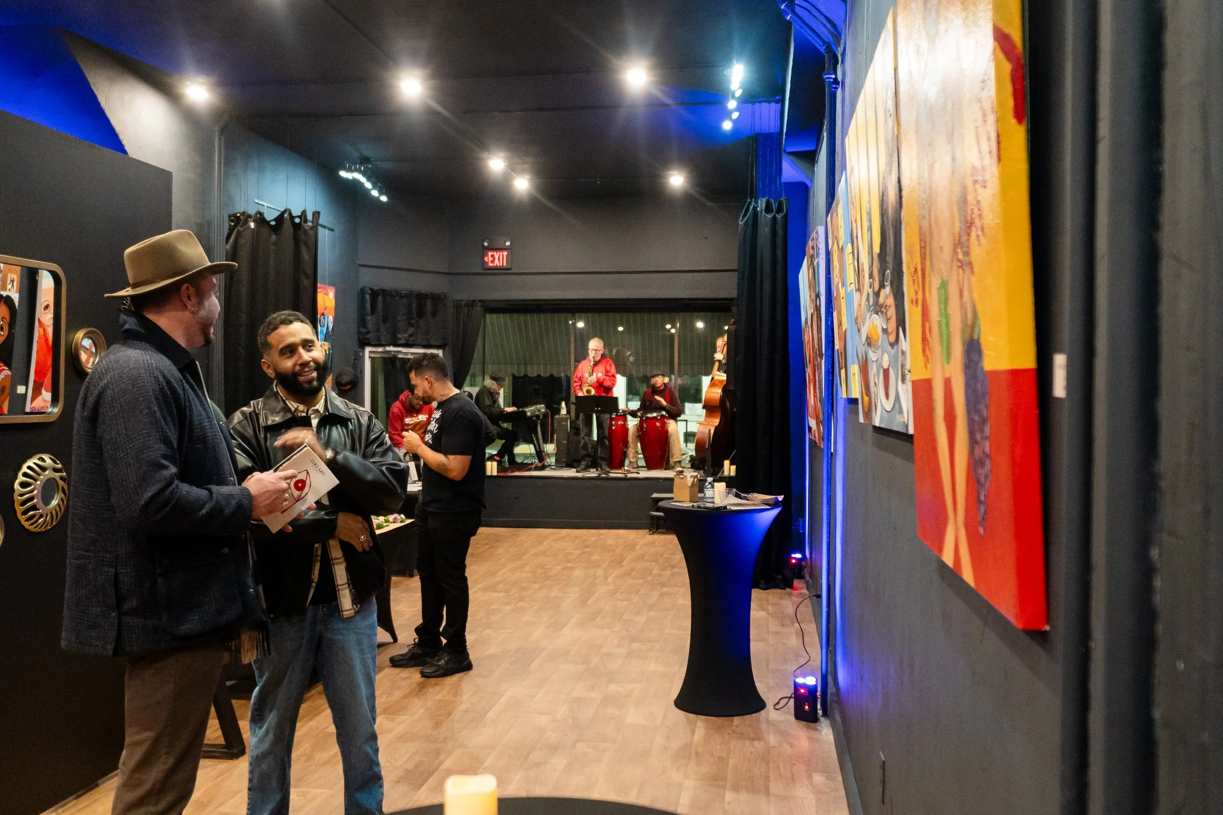 People socializing in an art gallery with paintings on the wall, a stage with musicians performing in the background, and a table with drinks.