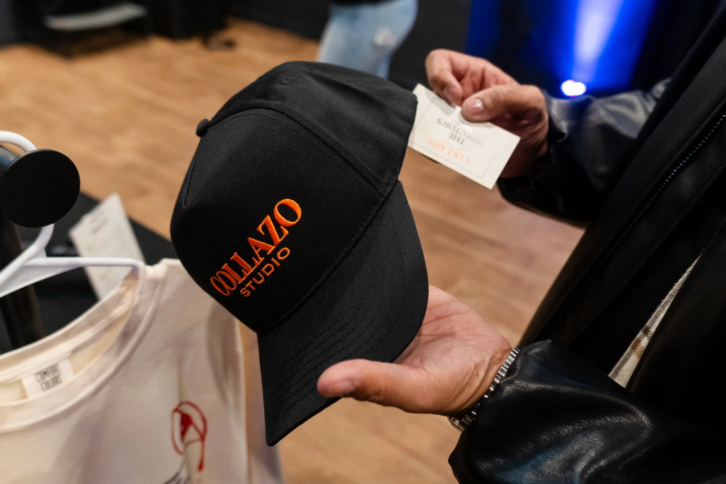Person holding a black cap with orange embroidered text that reads 'OZTARIO STUDIO', with a hand also holding a ticket and a clothing tag on a shopping bag nearby.