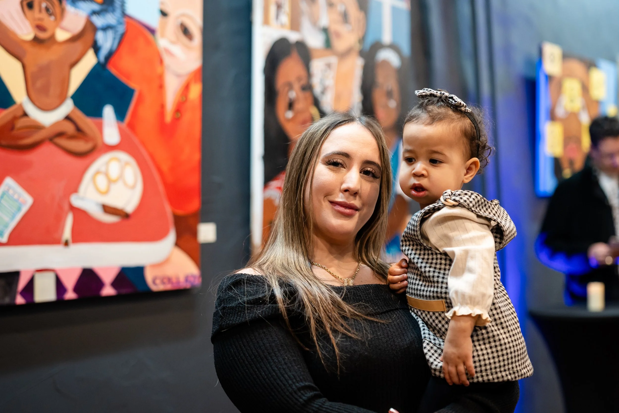 A woman holding a young girl in an art gallery with colorful paintings of children in the background.