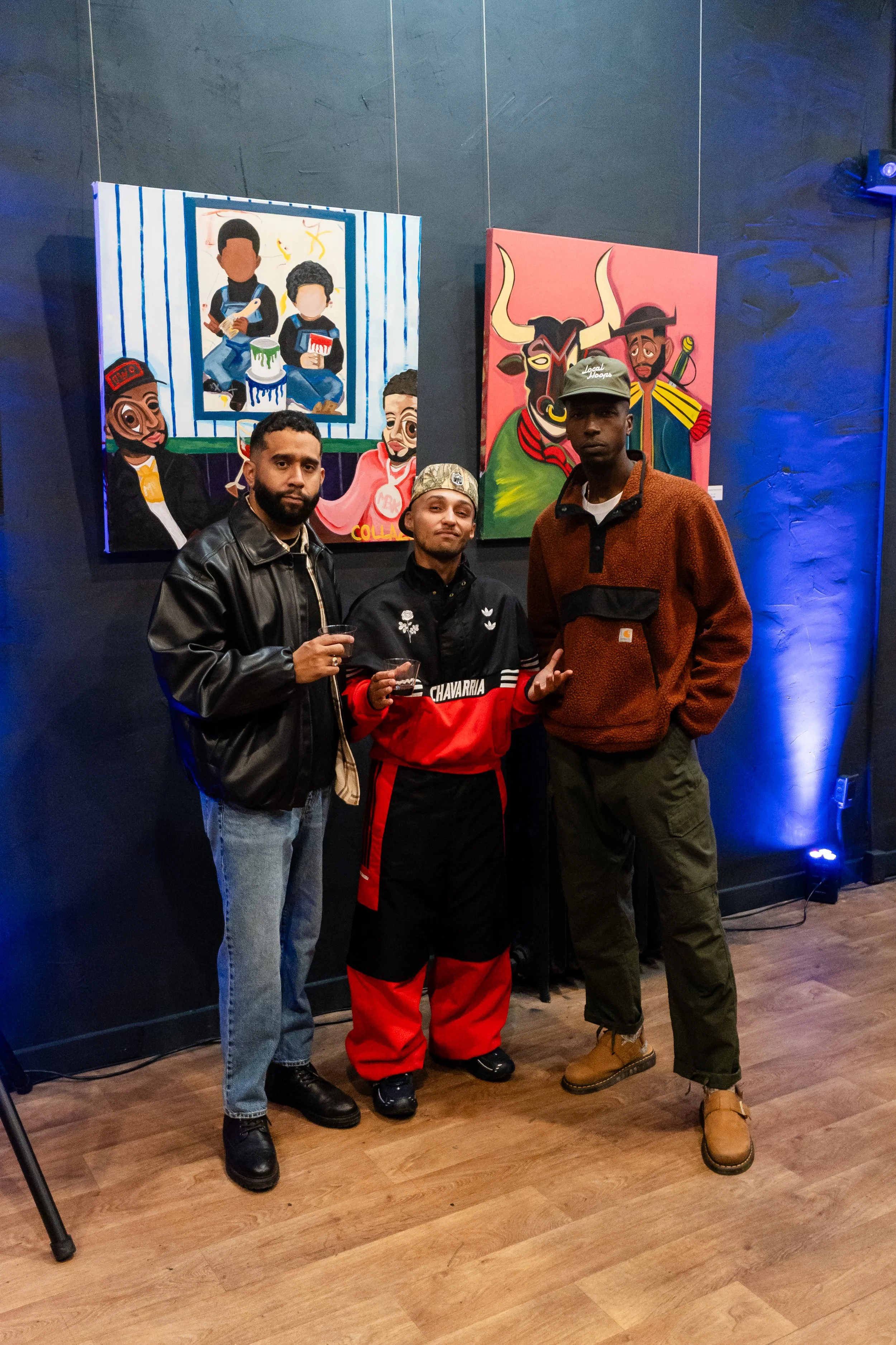Three men standing in front of colorful paintings on a dark wall at an art gallery or exhibition, with wooden flooring and blue lighting.