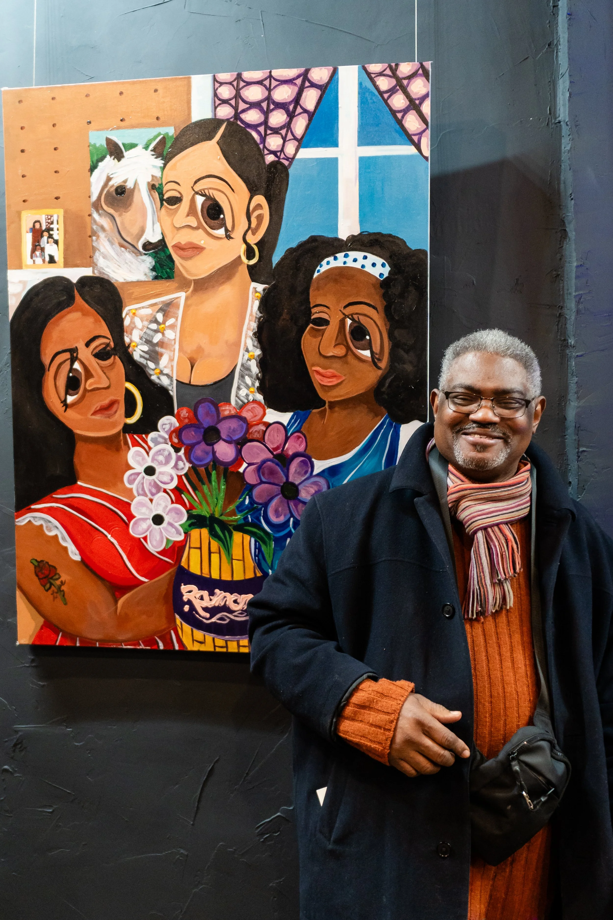 A man with glasses and gray hair smiling, dressed in a black coat, orange sweater, and striped scarf, standing in front of a colorful painting of four women with large eyes, holding flowers, inside an art gallery.