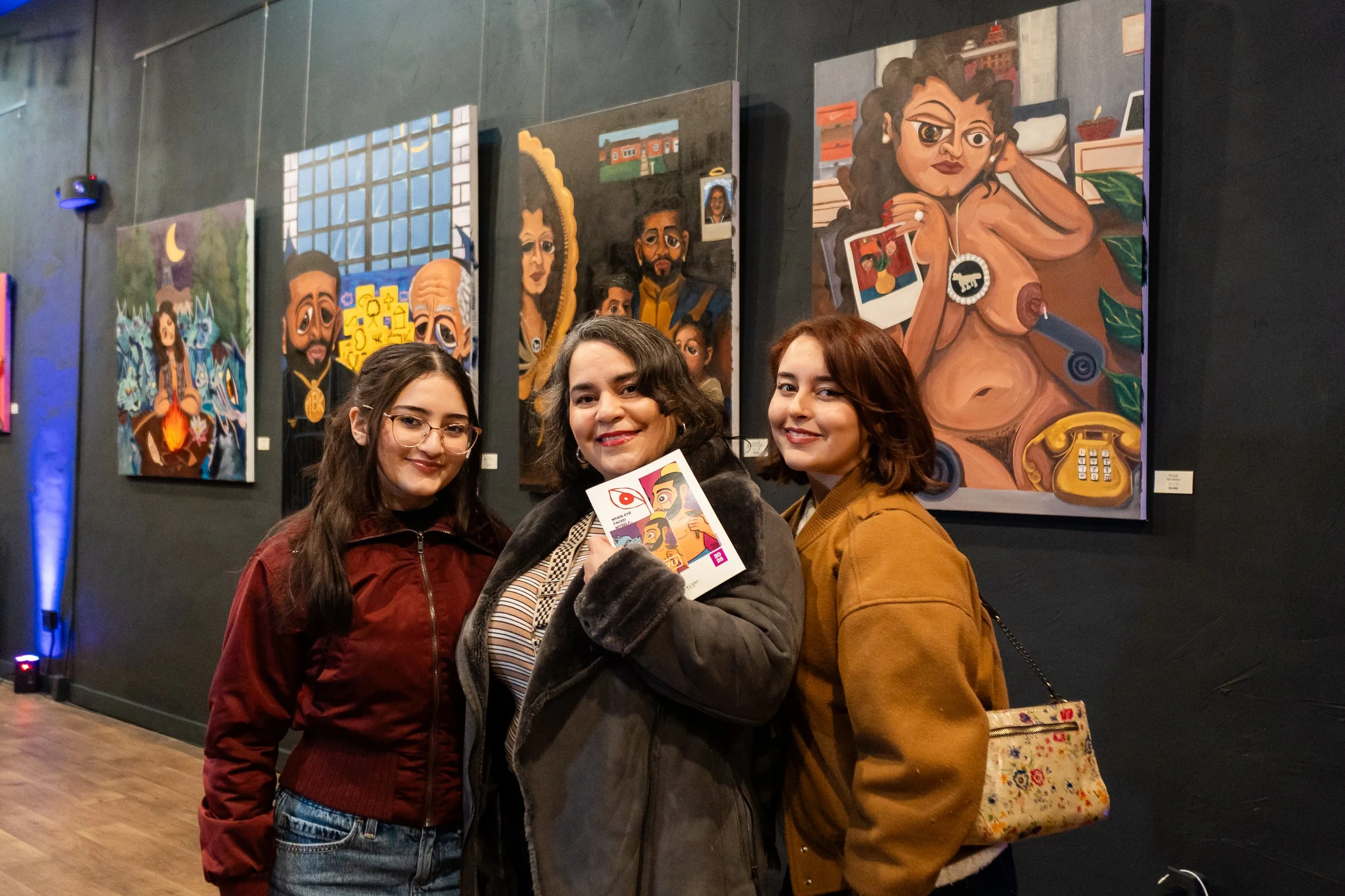 Three women standing close together and smiling in an art gallery, with colorful paintings displayed on a dark wall behind them.