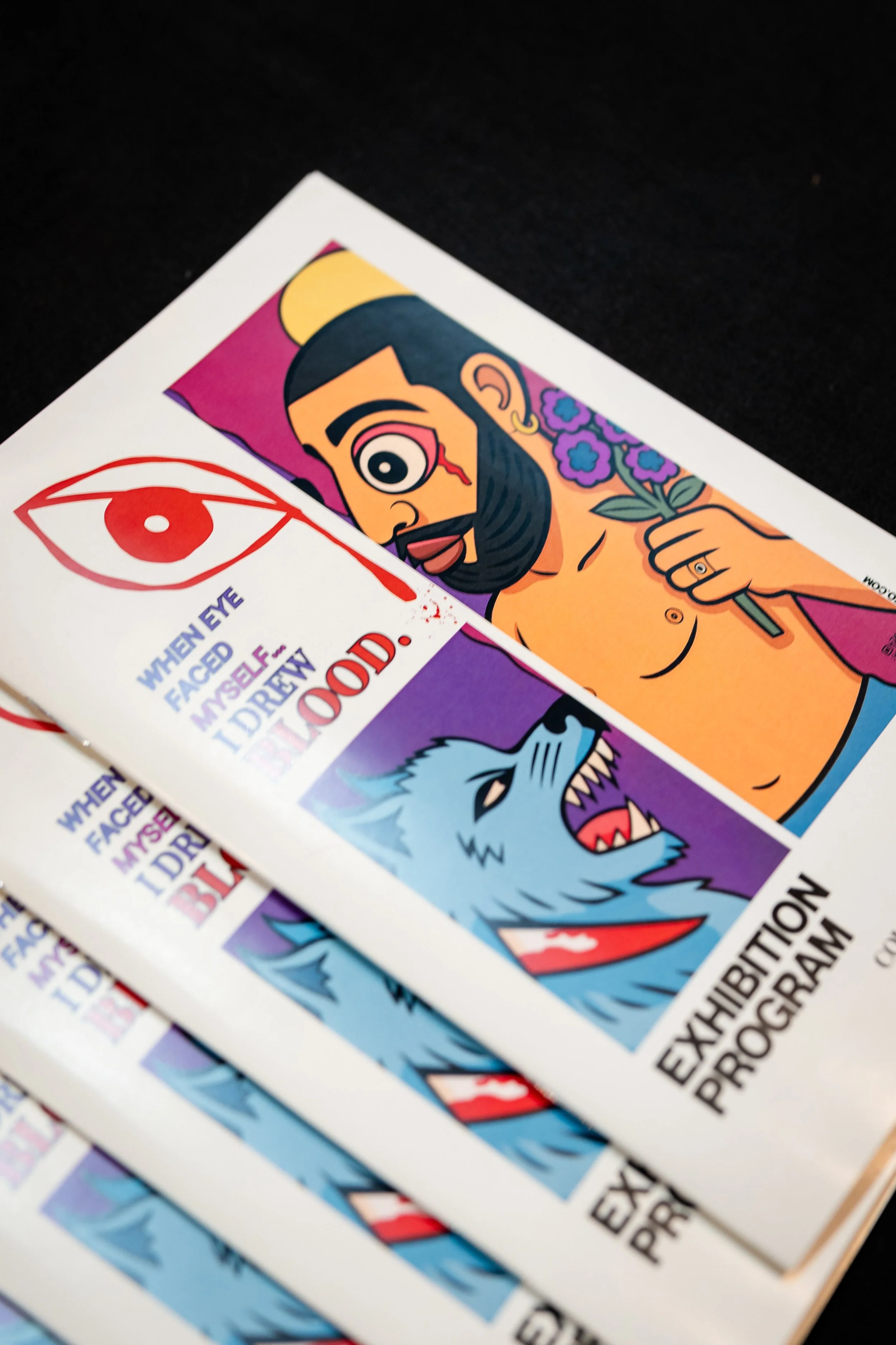 Multiple printed flyers for an exhibition with colorful artwork, including a cartoonish man holding flowers, a blue wolf, and text that reads 'When faced myself I drew blood. Exhibition Program'.