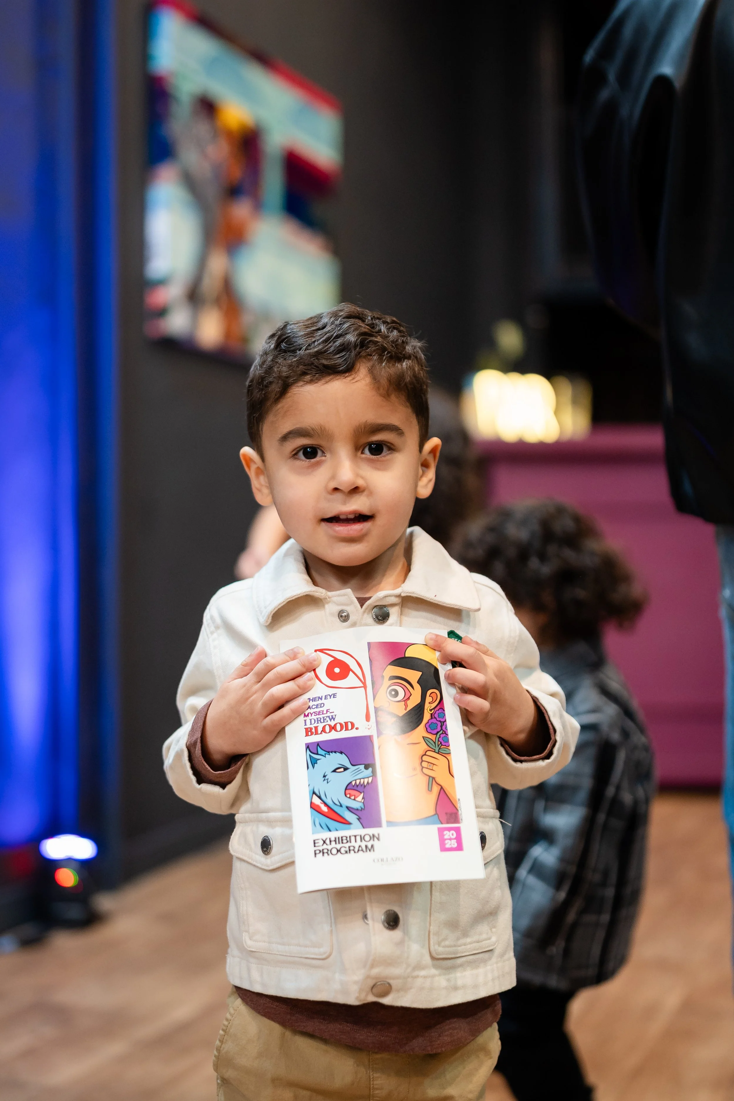 Young boy holding an exhibition program with colorful artwork, standing indoors among other children.