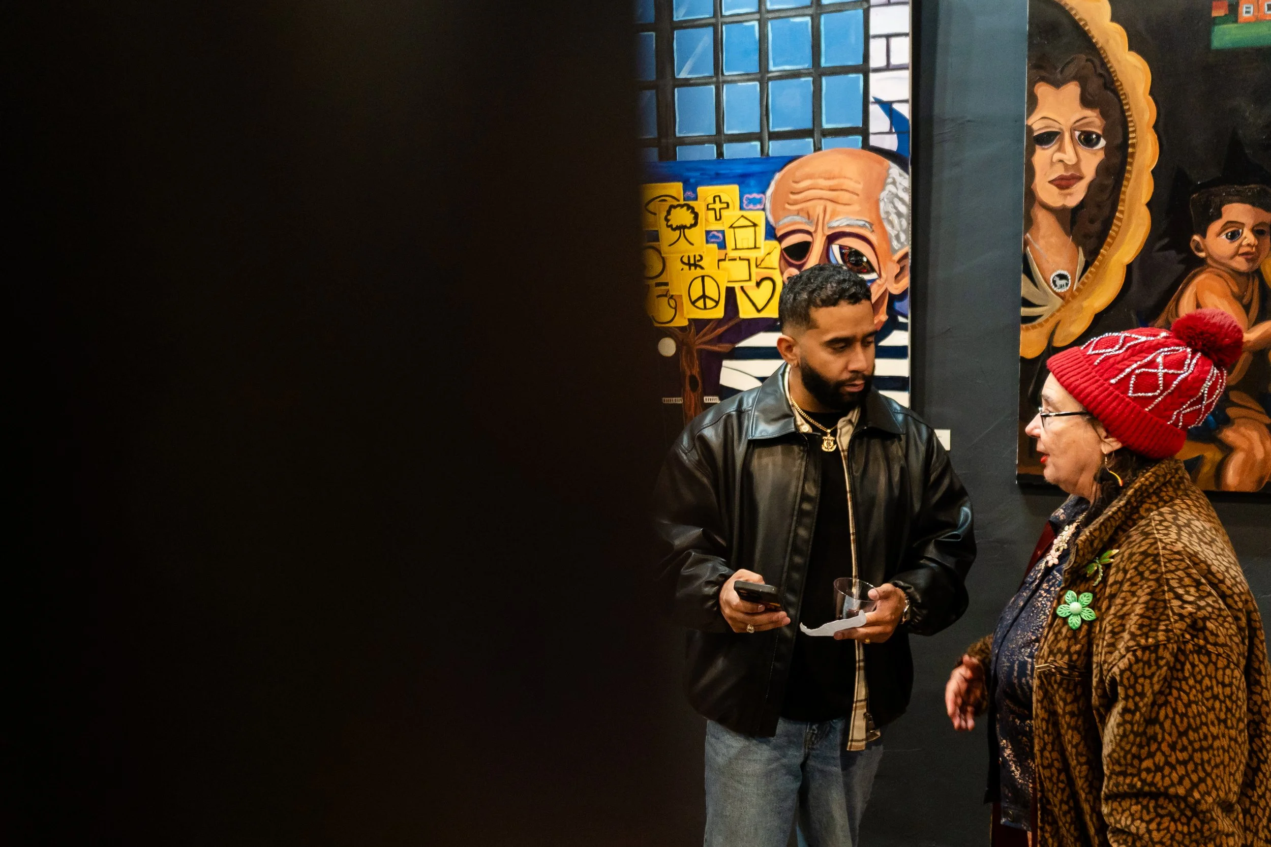Two people, a young man and an elderly woman, are engaged in conversation inside an art gallery. The young man is holding a smartphone and a glass, wearing a black leather jacket. The elderly woman is wearing a red knit hat, glasses, and a patterned 