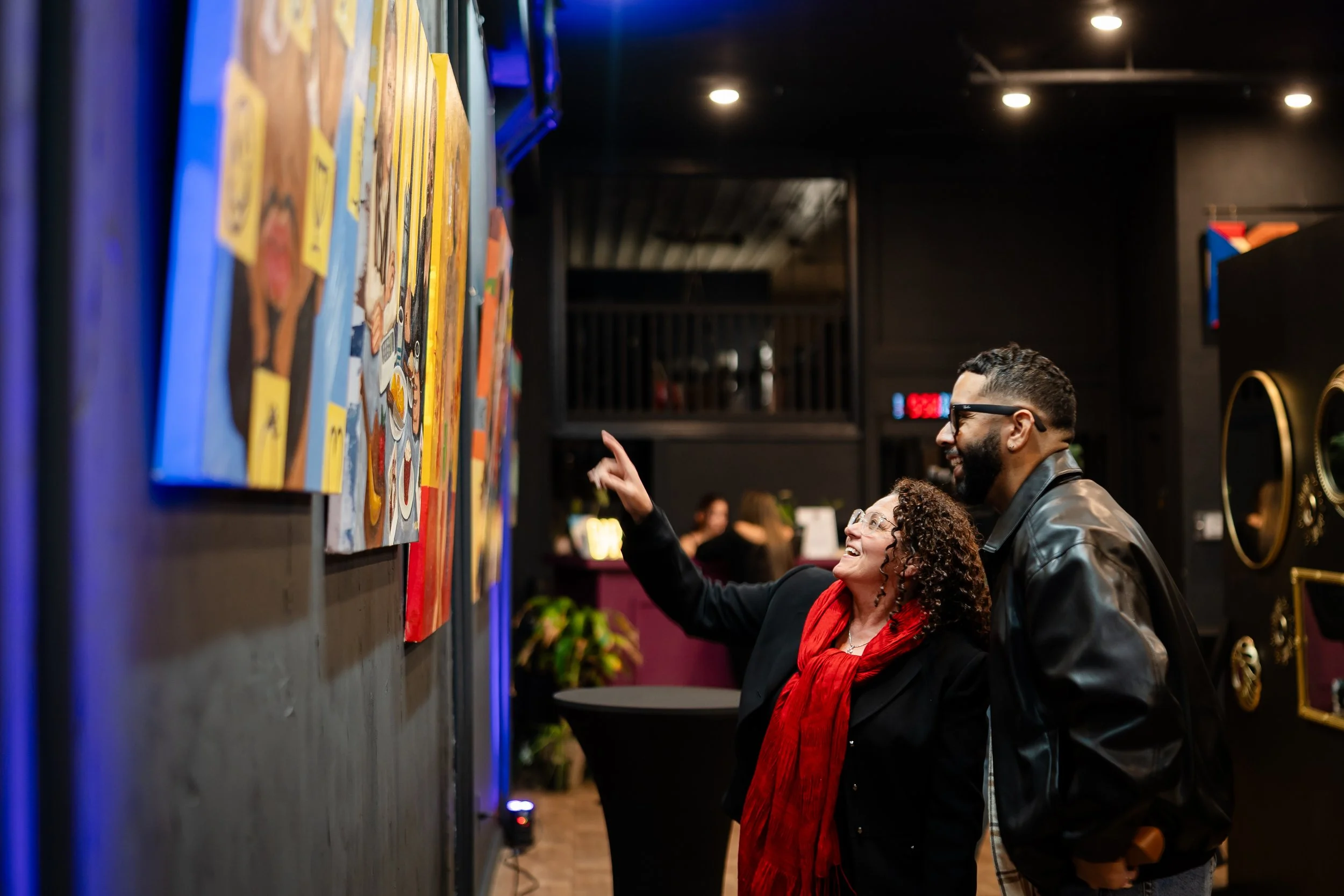 A woman and a man looking at colorful paintings on a gallery wall, smiling and talking. The woman is pointing at a painting, and the man is standing beside her, wearing glasses and a leather jacket.