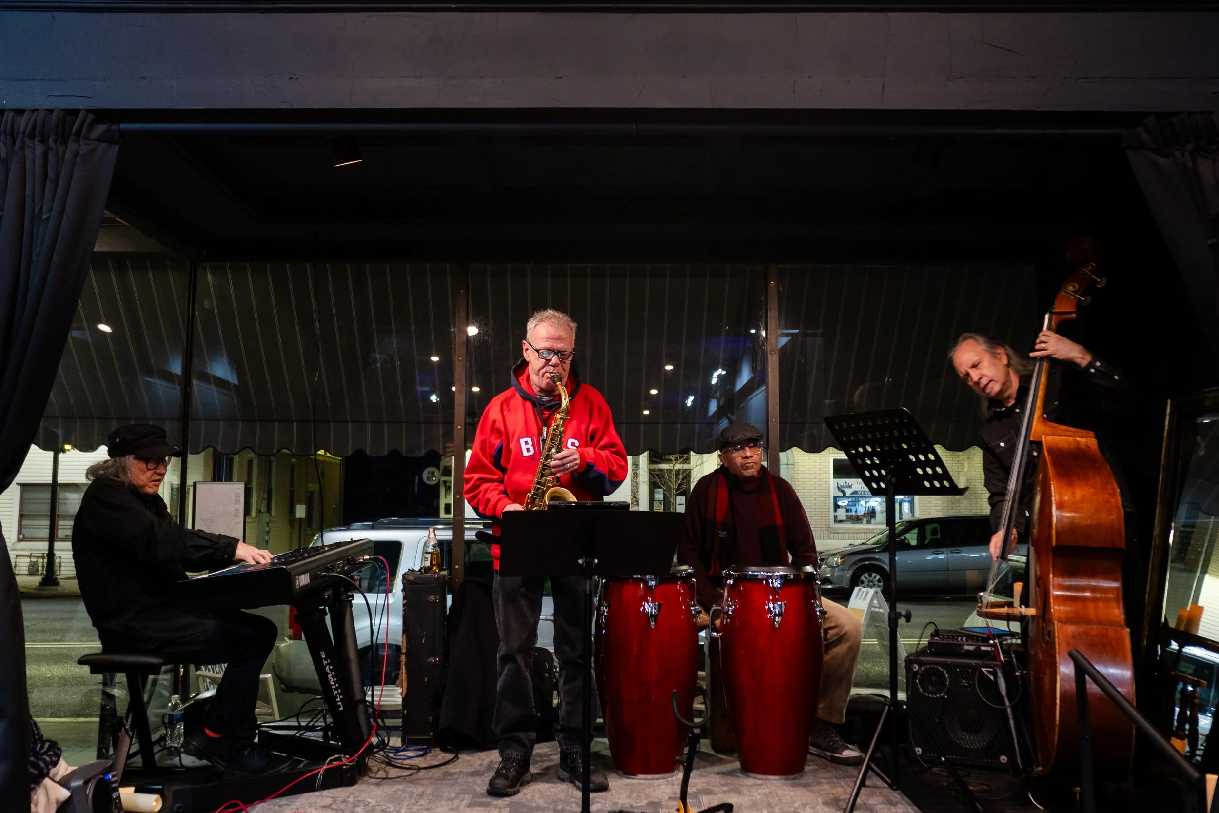 Four musicians performing live on stage: a keyboard player on the left, a saxophonist in the center, a percussionist with conga drums next to the saxophonist, and a double bass player on the right.