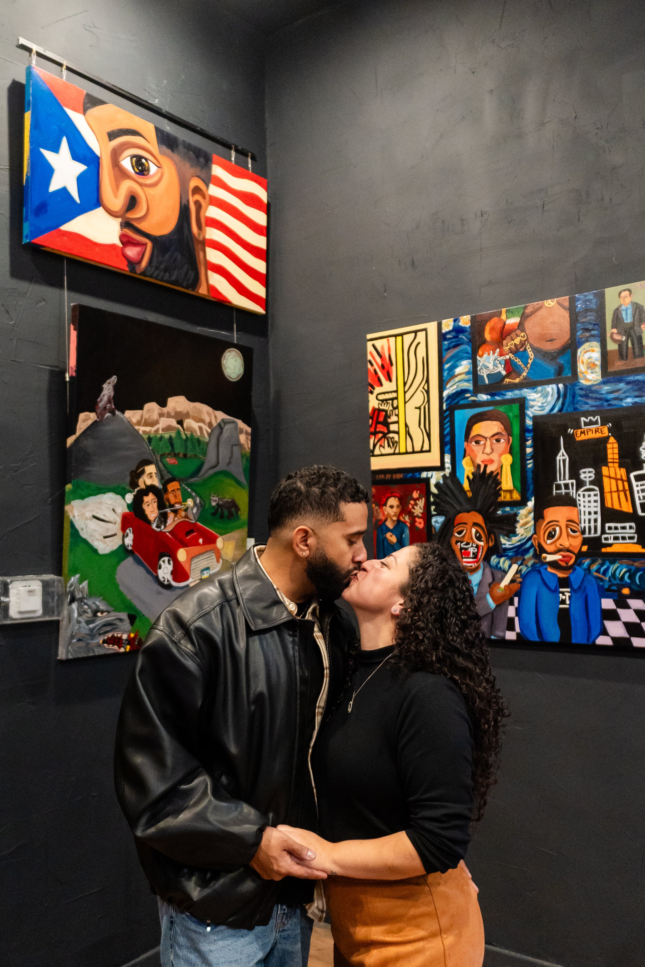 A couple kissing in an art gallery with colorful paintings on the dark wall behind them.