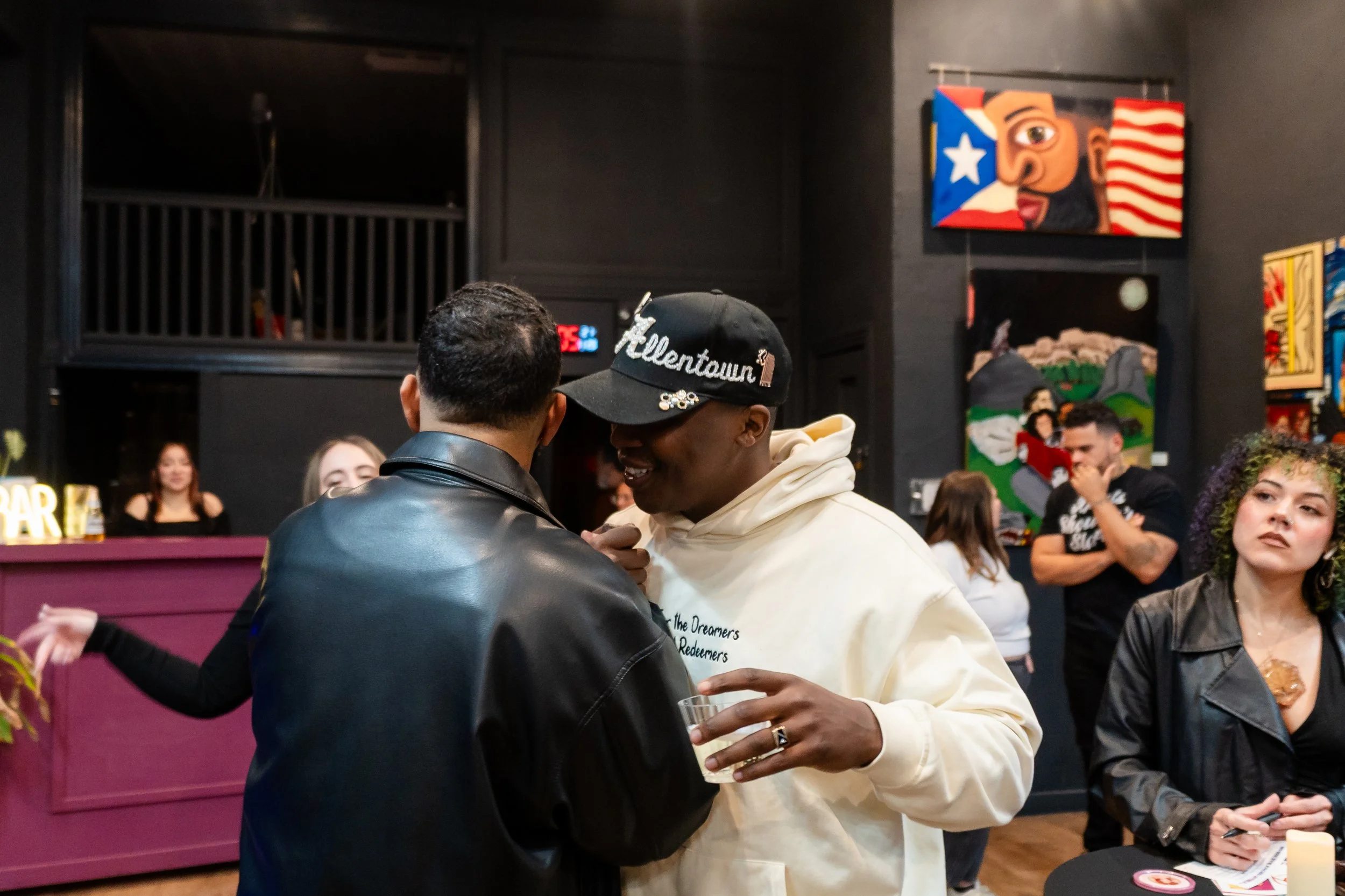 Two men are talking and smiling, one with a black leather jacket and the other with a cream hoodie and a black cap that reads 'Helltown'. In the background, other people are socializing in a room with colorful artwork on dark walls.