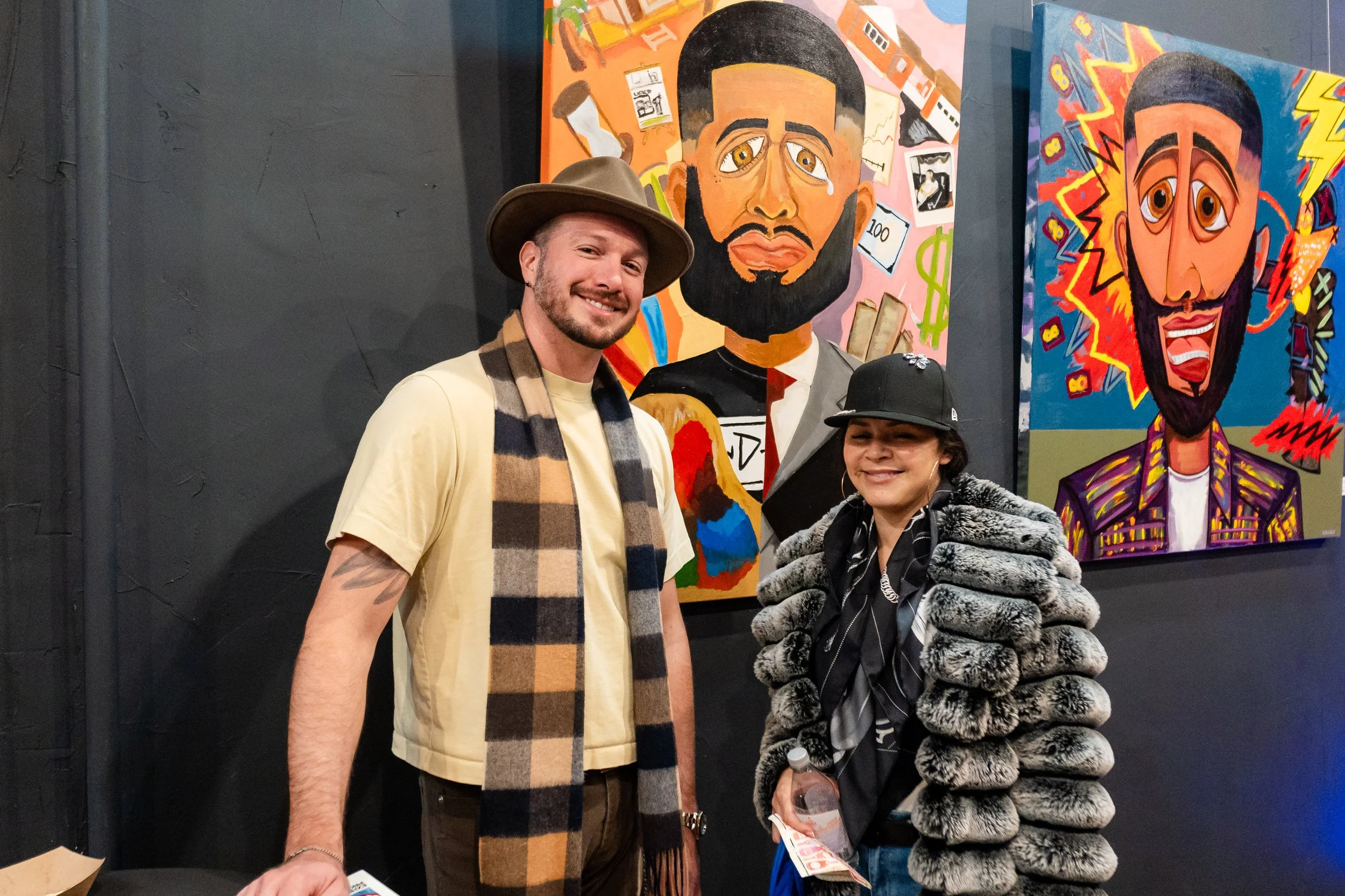 Two people smiling at an art gallery, standing in front of colorful paintings of stylized faces on a dark wall.