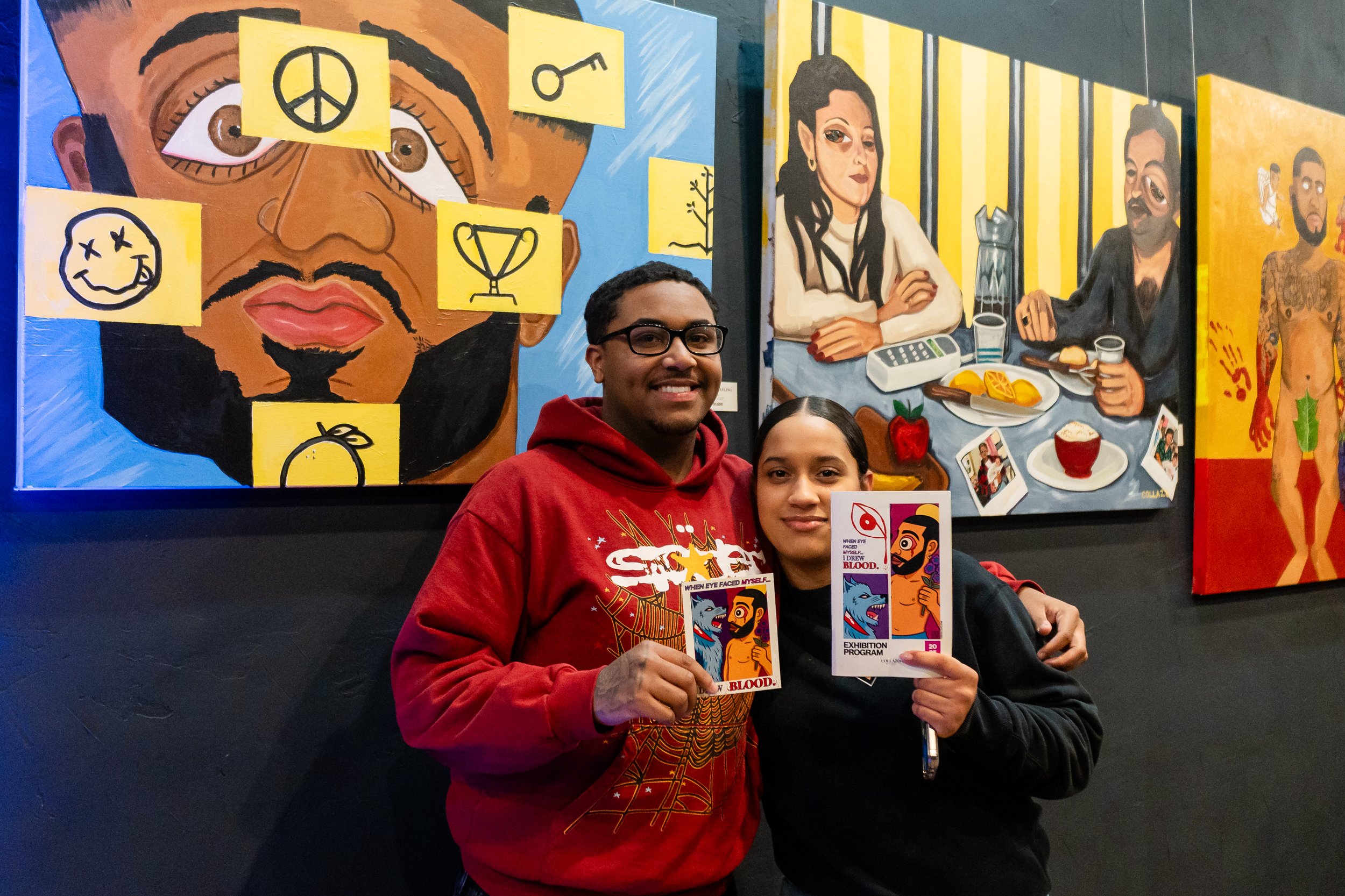 A man and woman standing in front of colorful artwork at an exhibition, holding event programs that show artistic illustrations of faces and blood.