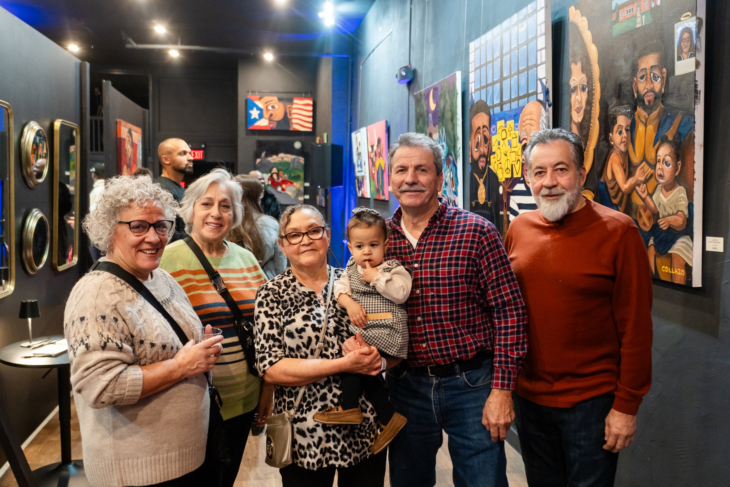 A group of six people, three women, two men, and a young child, standing inside an art gallery with colorful paintings on the walls. They are smiling at the camera.