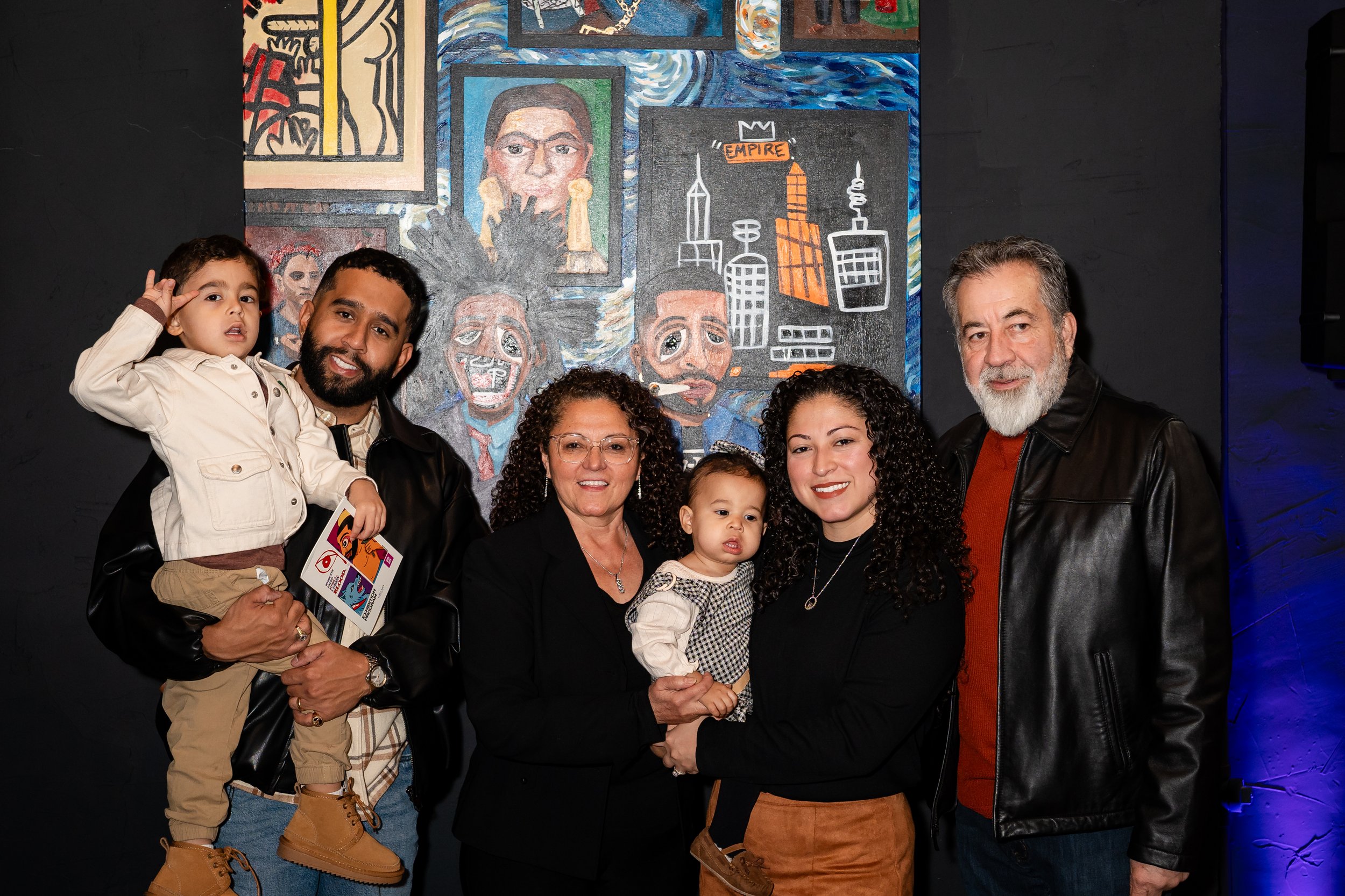 Group of six people, including two children, smiling and posing in front of a colorful artistic mural on a black wall. The mural features abstract portraits and cityscape drawings.