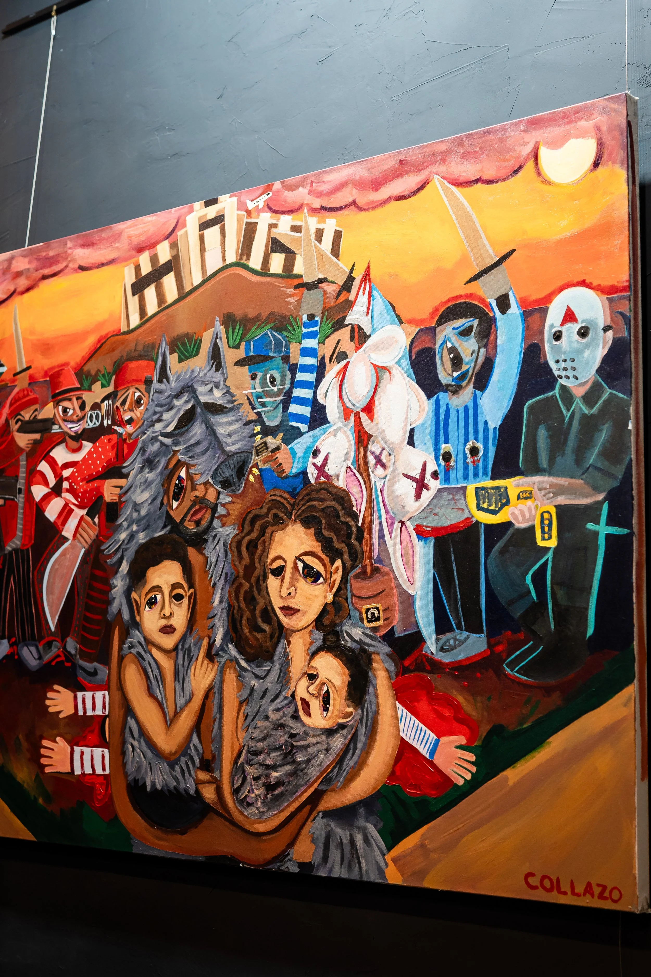 Colorful painting depicting a chaotic scene with people in costumes, some wearing masks and animal elements, holding weapons, and appearing distressed. The background shows a sunset with a large moon, and a hillside with trees and a structure, possib