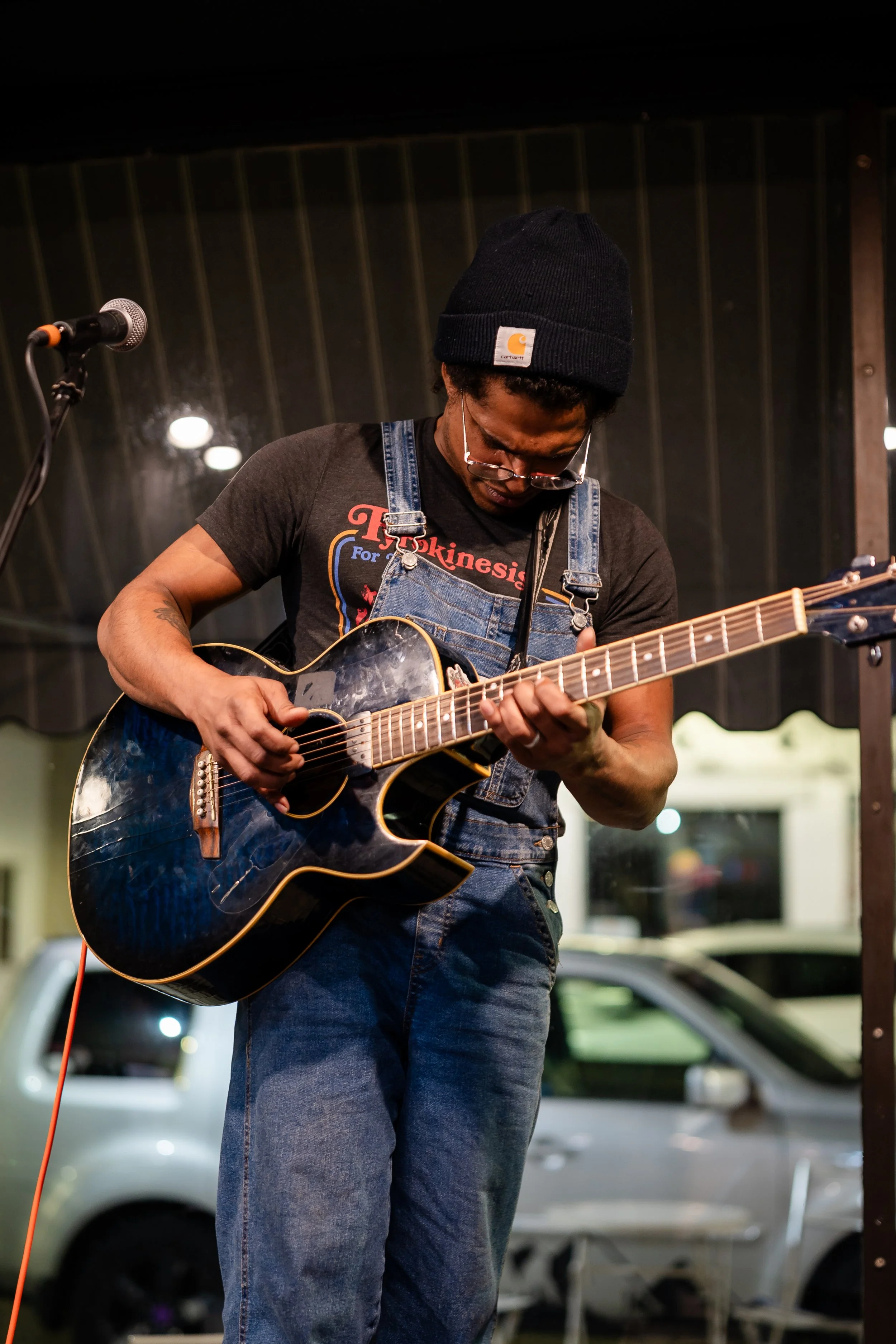 A man wearing a black Carhartt beanie, glasses, a black T-shirt with red and blue text, and denim overalls playing an acoustic guitar on stage.