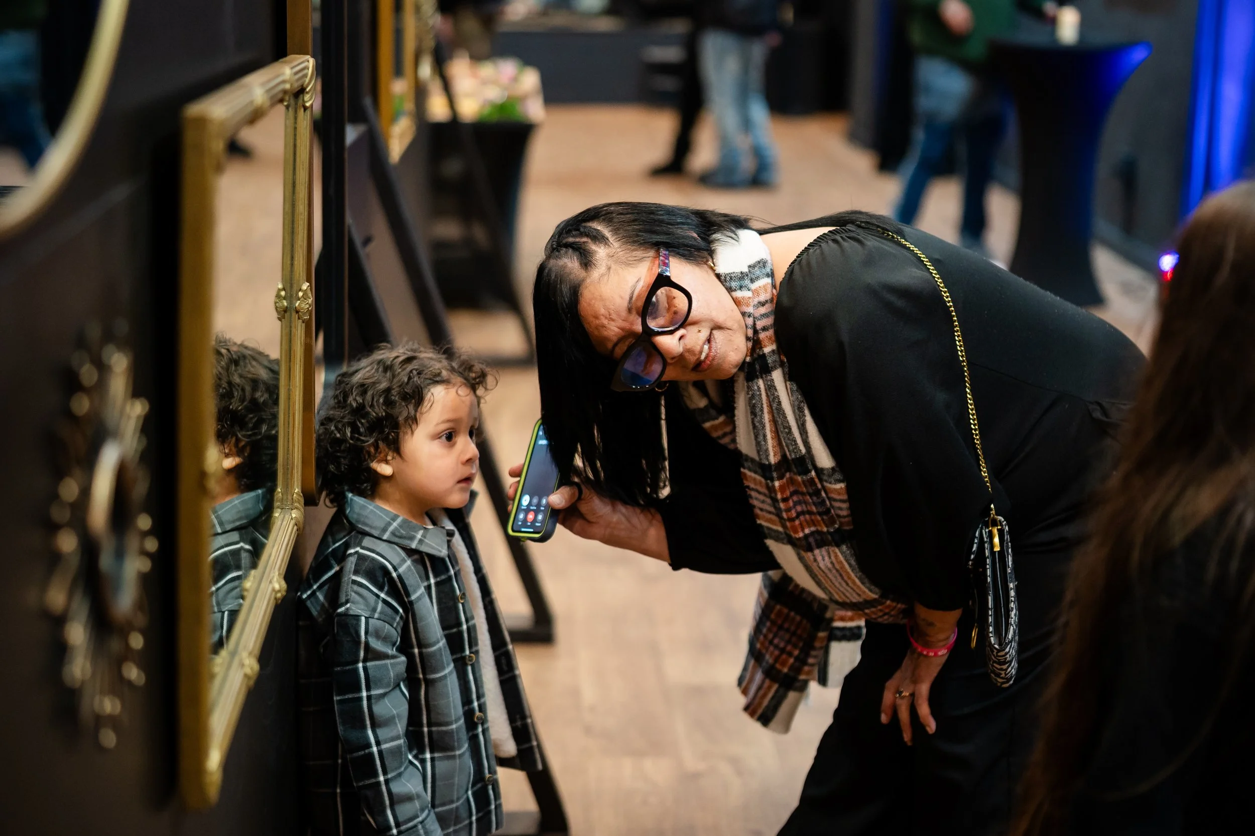 A woman with glasses and a scarf is talking to a young boy with curly hair, wearing a plaid jacket, in an indoor art gallery. The woman is holding a smartphone.