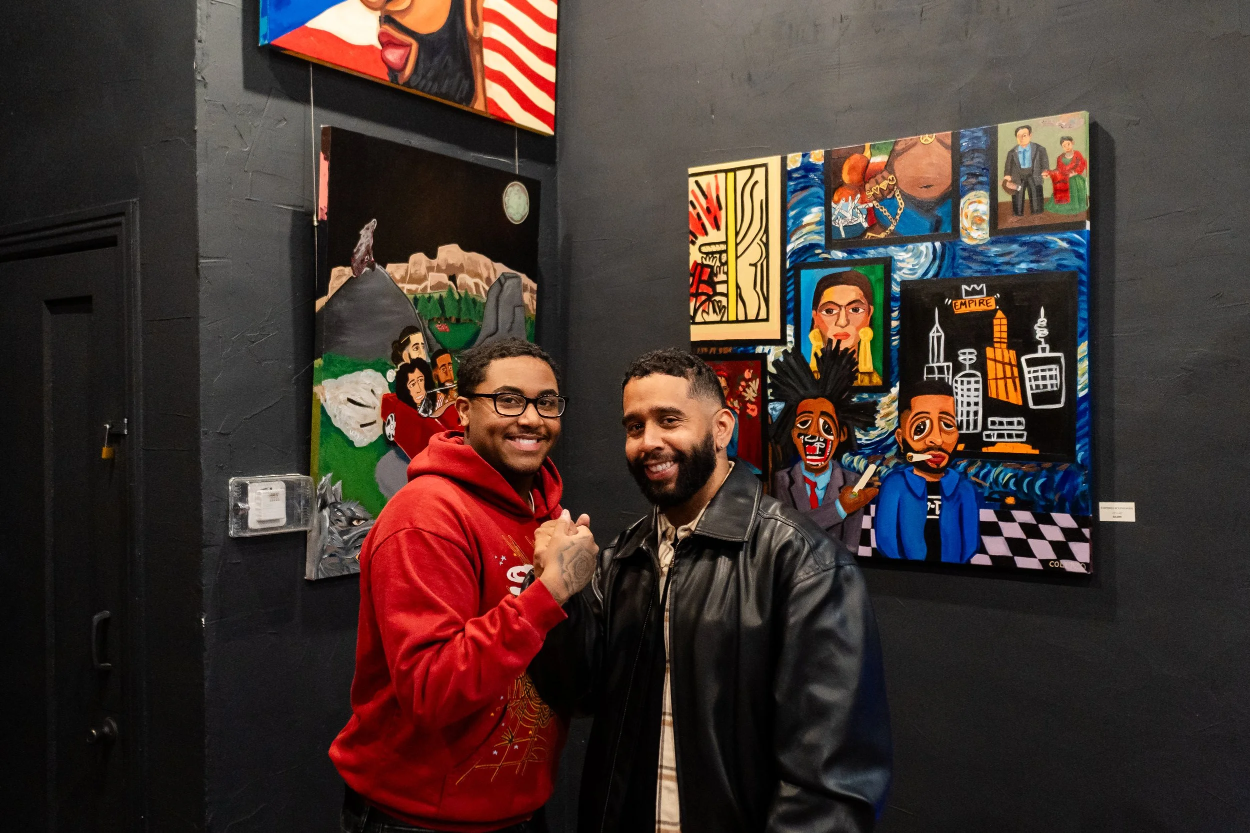 Two men smiling and shaking hands in front of colorful artwork in a dark gallery.