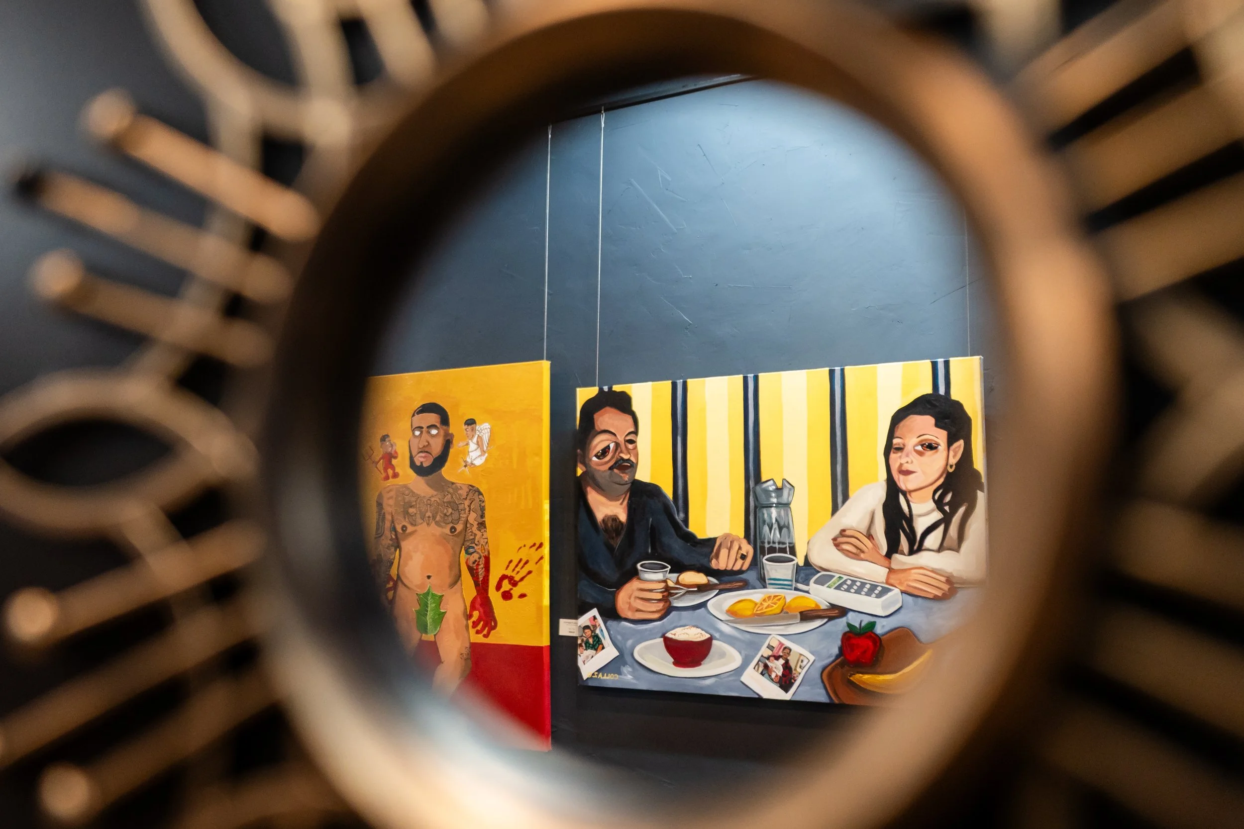 View of colorful paintings hanging on a dark wall seen through a circular object, with one painting depicting two people at a table and another showing a tattooed man with a green leaf pubic area.