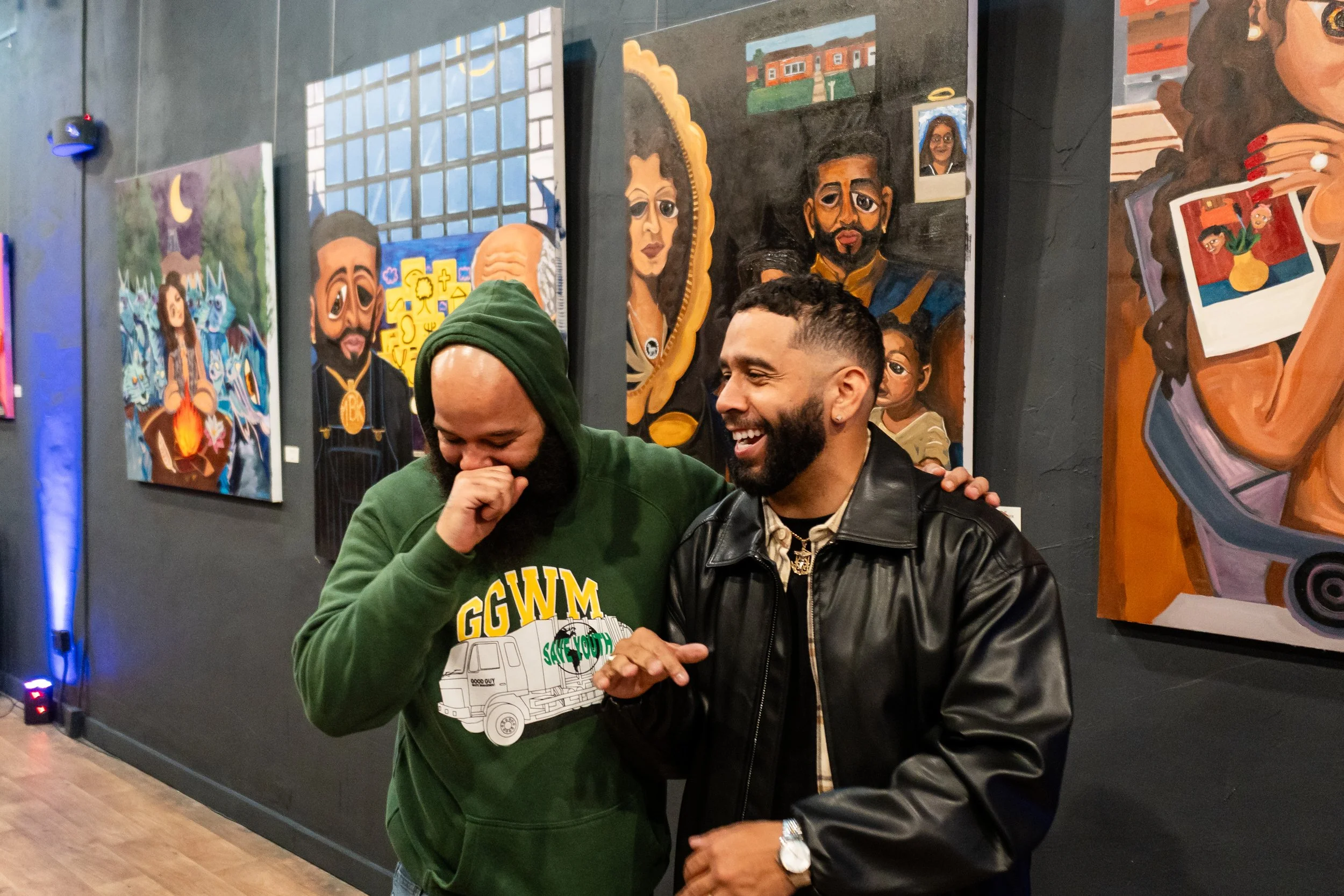 Two men are standing together, smiling and laughing in an art gallery with colorful paintings on the wall behind them. One man has a beard and is wearing a black leather jacket; the other is wearing a green hoodie with a beanie.