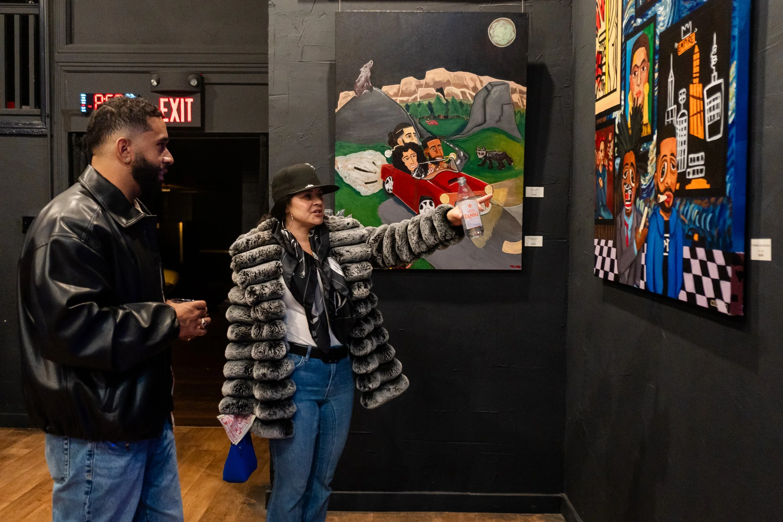 Two people are viewing colorful paintings in an art gallery. The woman is pointing at one of the paintings, which depicts a scenic outdoor landscape with cartoon-like characters in a red convertible. The man is standing next to her, observing the art