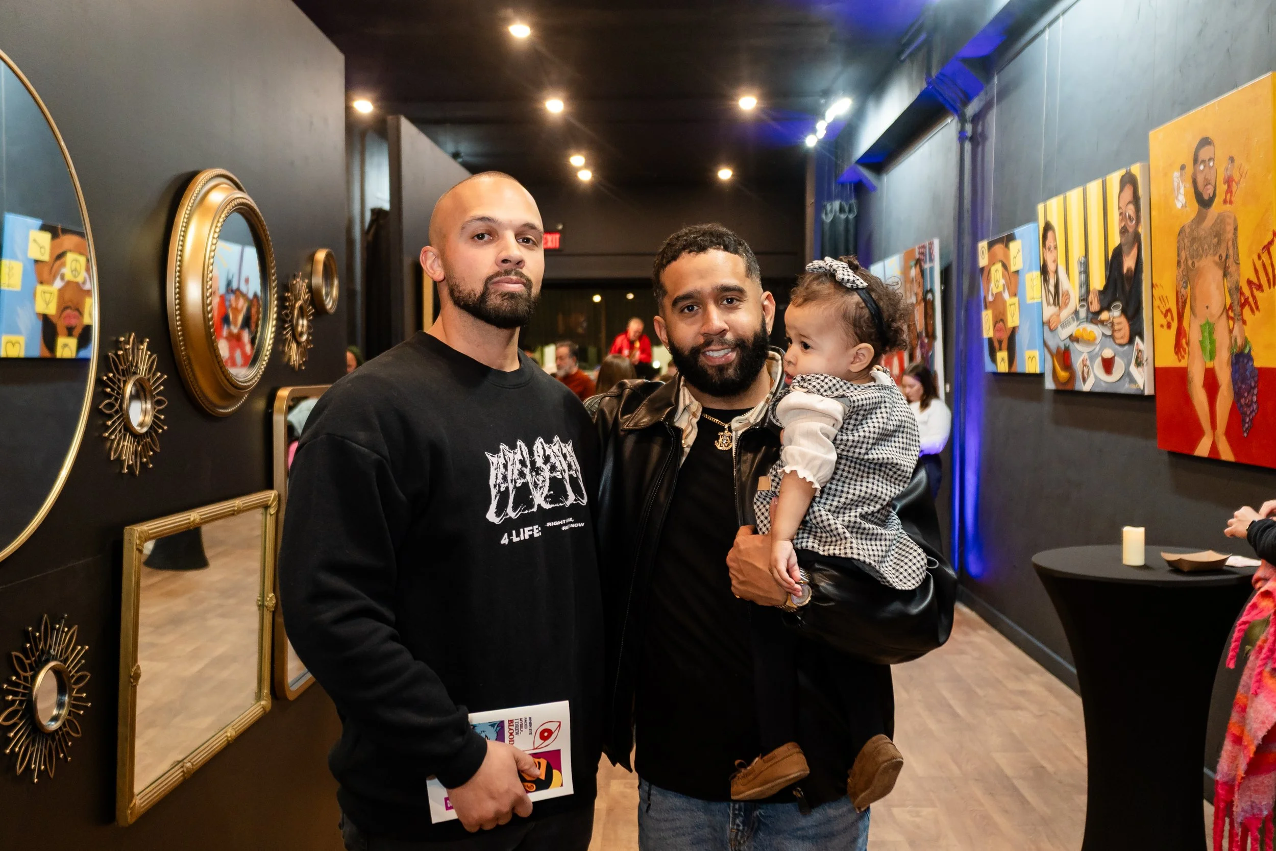 Two men standing with a young child in an art gallery or exhibition space with colorful paintings and mirrors on the wall.
