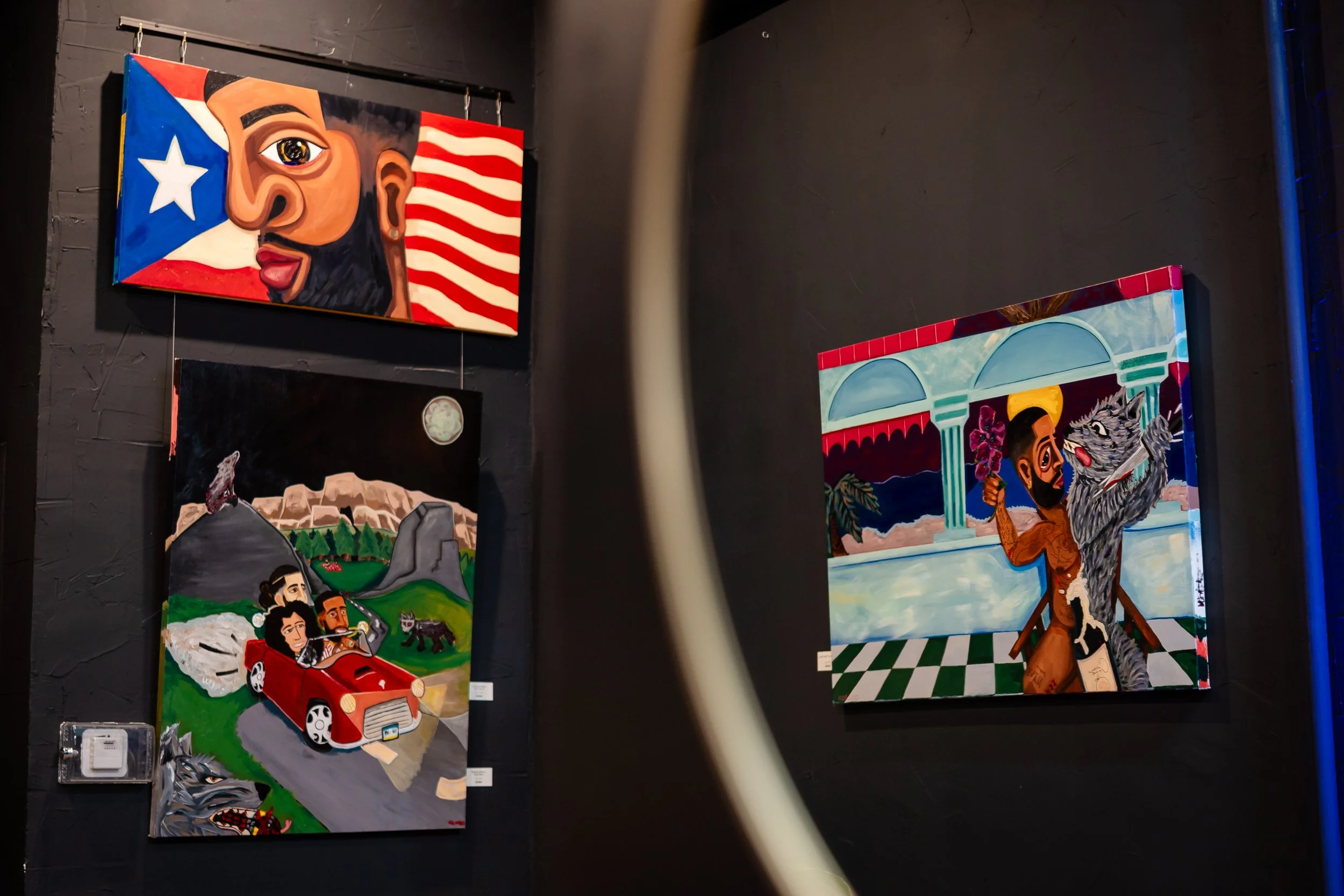 Three colorful paintings on a dark wall. The top left painting depicts a stylized portrait of a person with a beard, an eye, and an earring, over a Texas flag background. The bottom left painting shows a couple driving a red car on a rural road with 