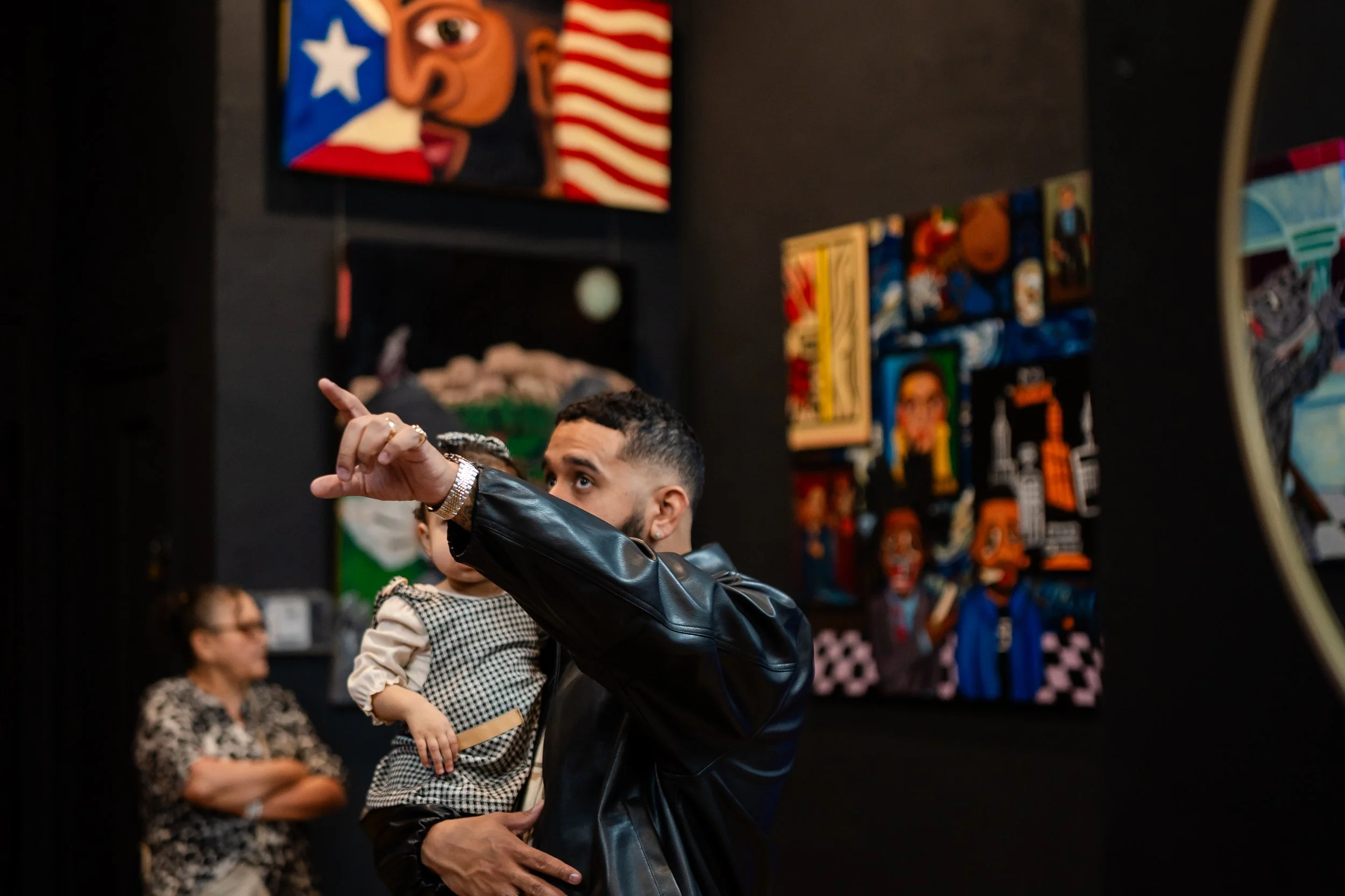 A man holding a child at an art gallery, pointing towards artwork on display with abstract paintings on dark walls behind them.