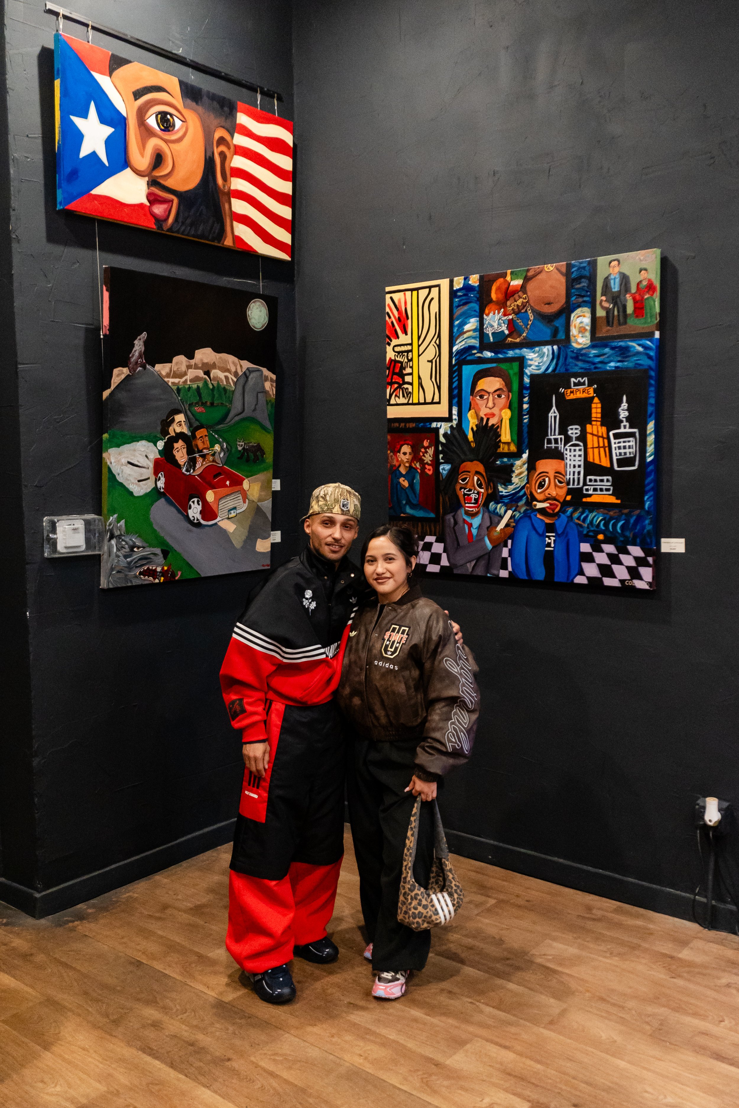 Two people standing and smiling in an art gallery with colorful paintings on dark walls.