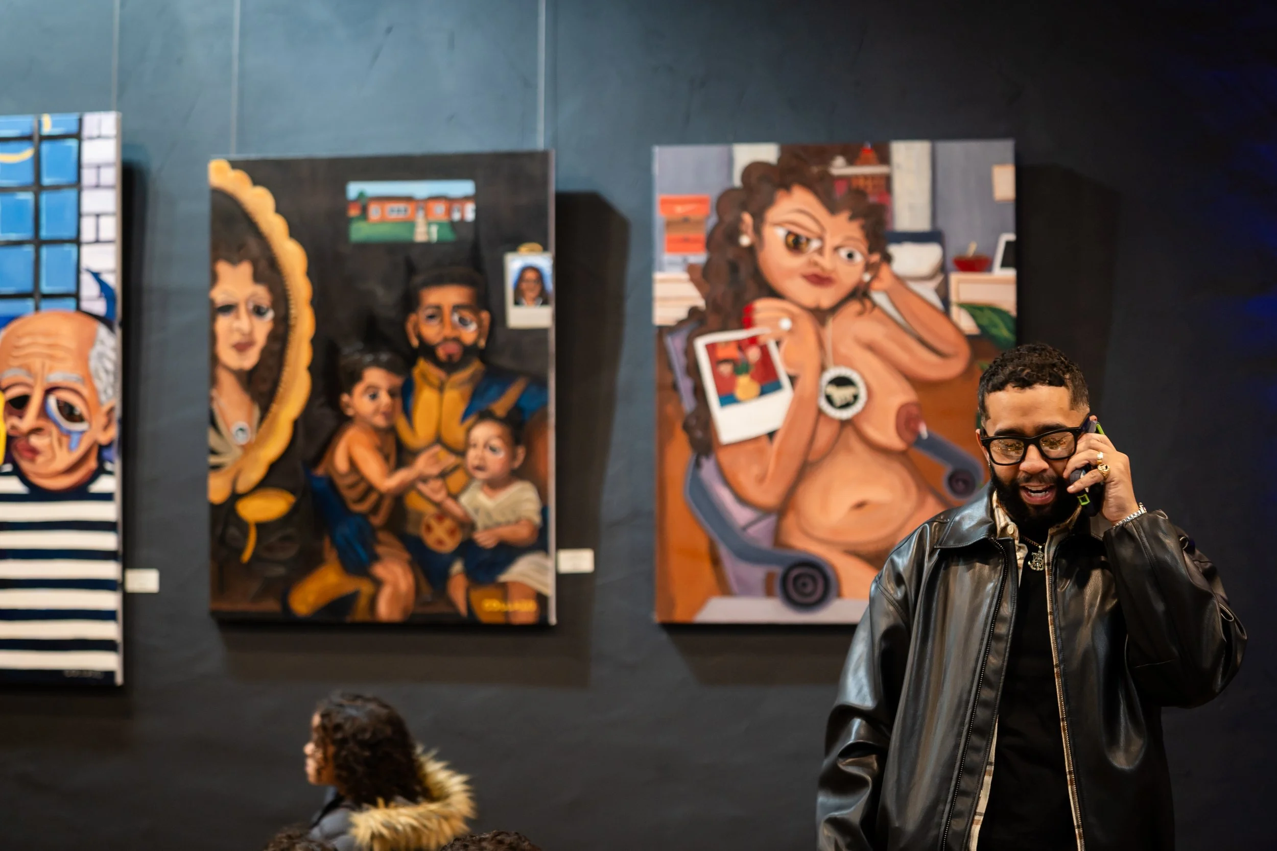 A man with glasses and a black leather jacket talking on a cell phone in an art gallery, with colorful paintings on the wall behind him, including a painting of a woman with curly hair holding a Polaroid camera and paintings of families.