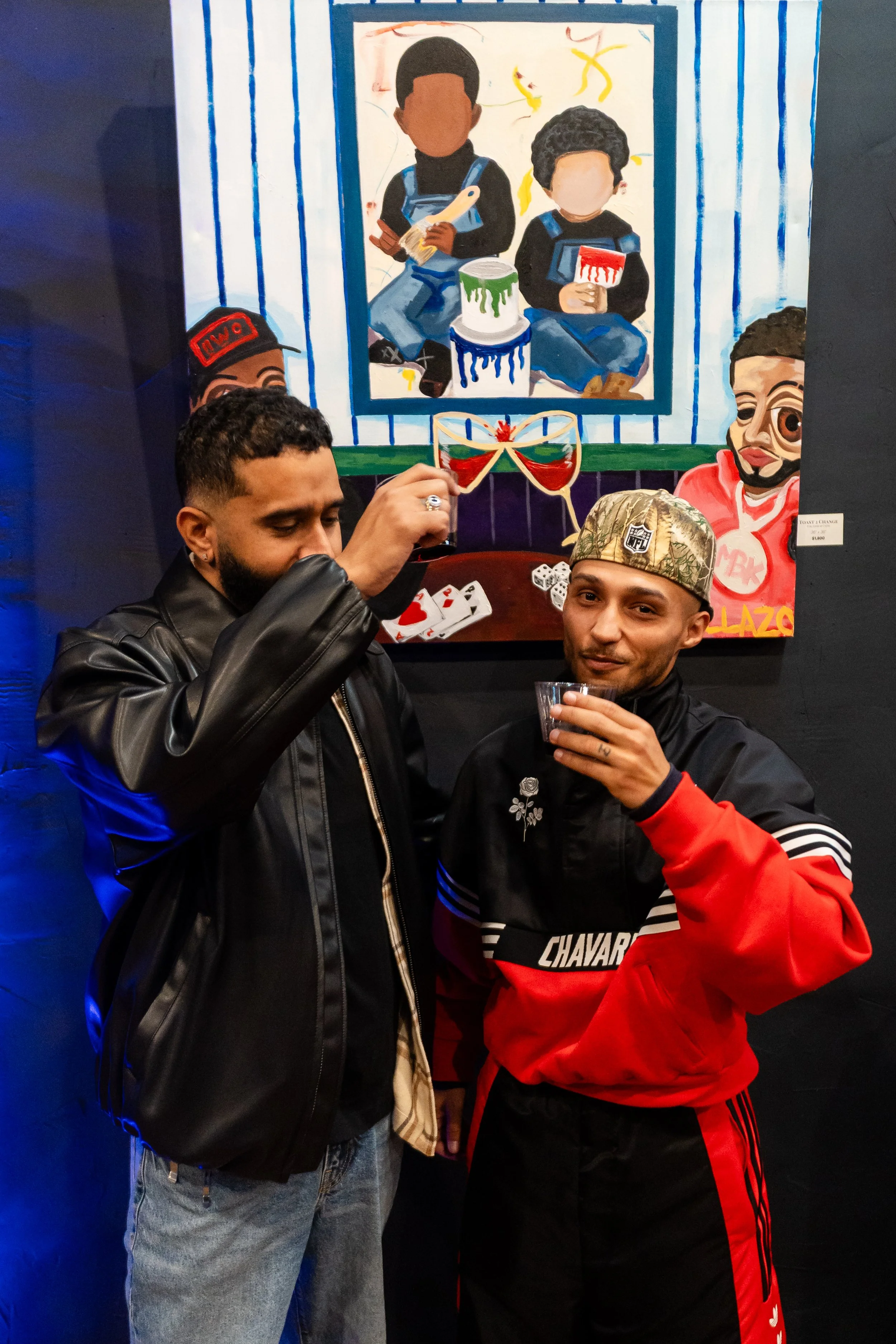 Two men standing in front of a colorful painting, one wearing a black leather jacket and the other a red and black sports jacket with a cap, raising glasses in a toast.