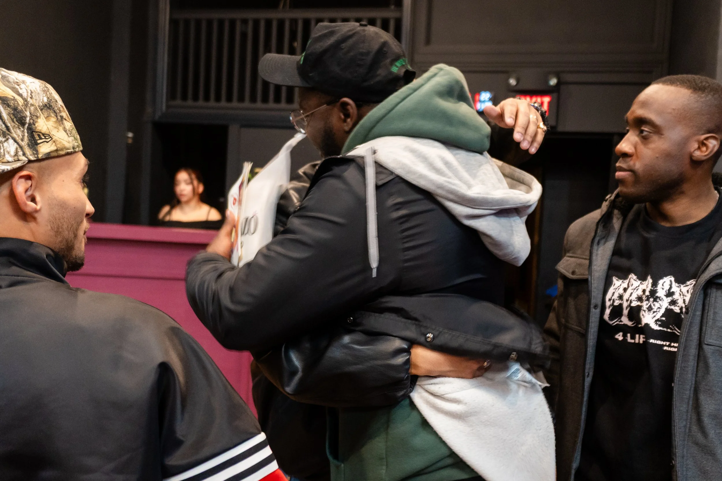 Three men in casual clothing engaged in conversation in an indoor setting, with a woman at a reception desk in the background.